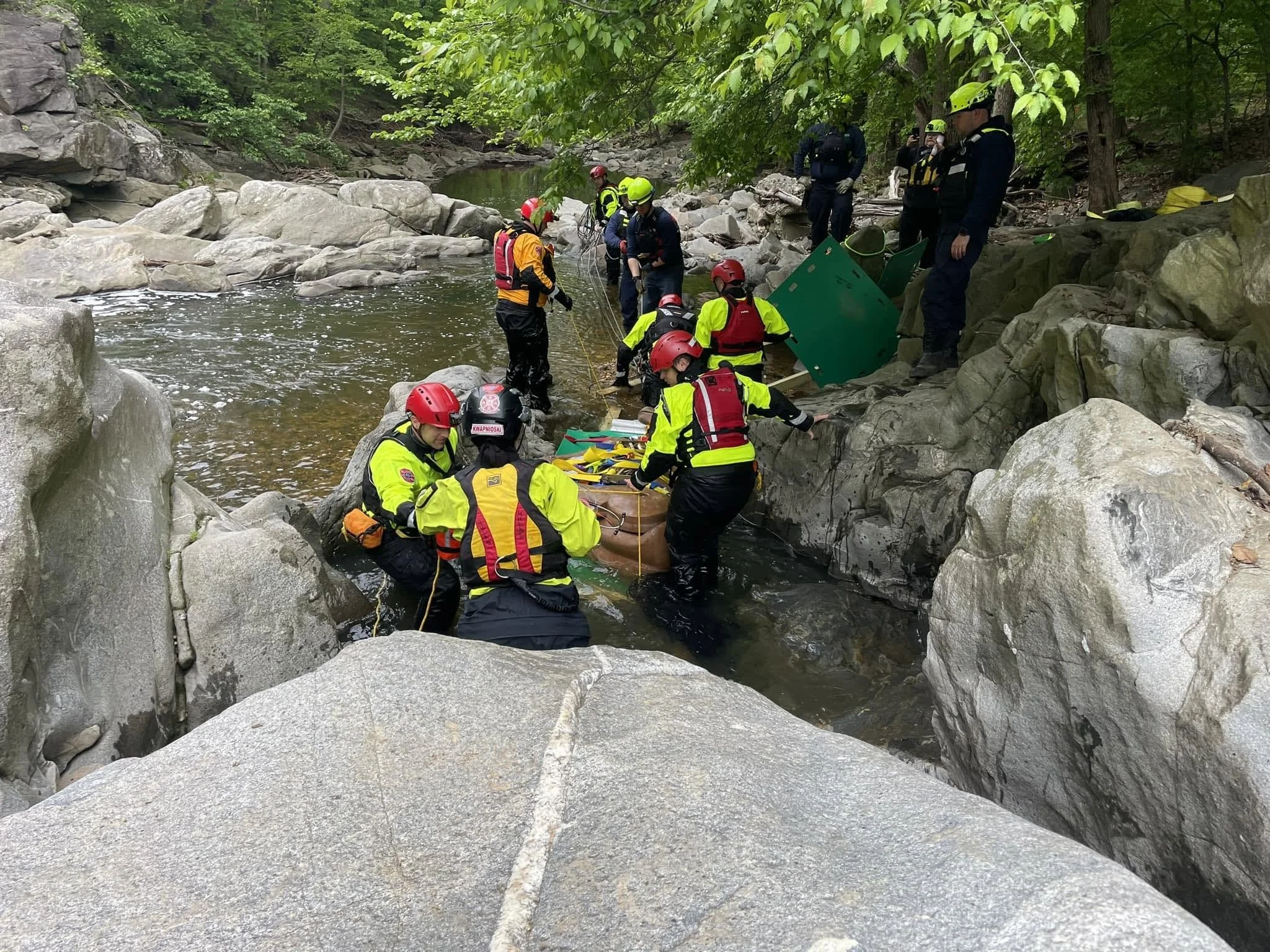 Rescue team members in helmets and life jackets working together at the edge of a rocky river in a forested area, preparing for rescue or training activity.