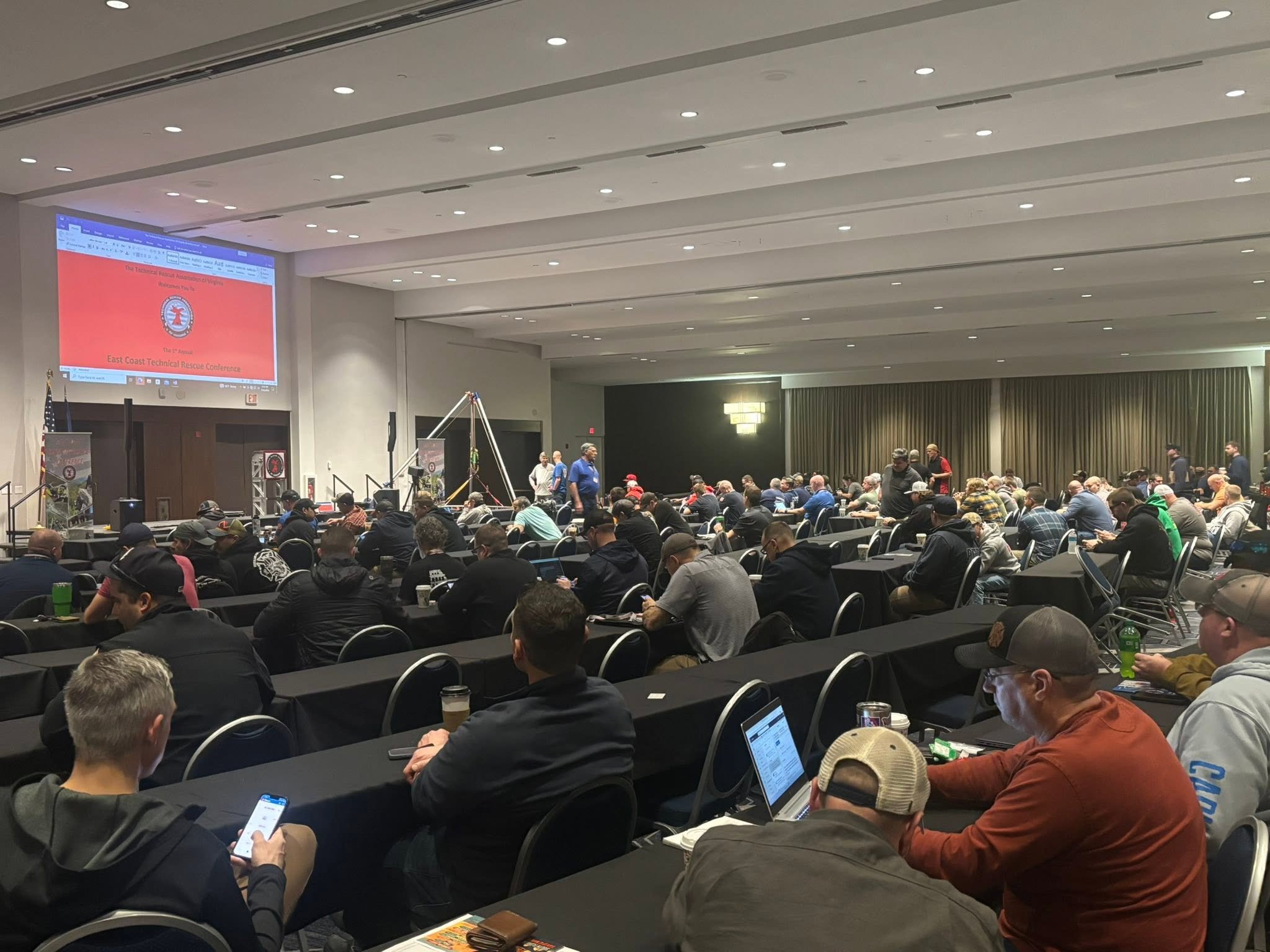 A large conference room filled with people sitting at long tables, some looking at laptops or phones. A large screen displays the East Coast Technical Rescue Conference logo and title. The room has beige walls, a high ceiling with recessed lighting, and curtains along one wall.