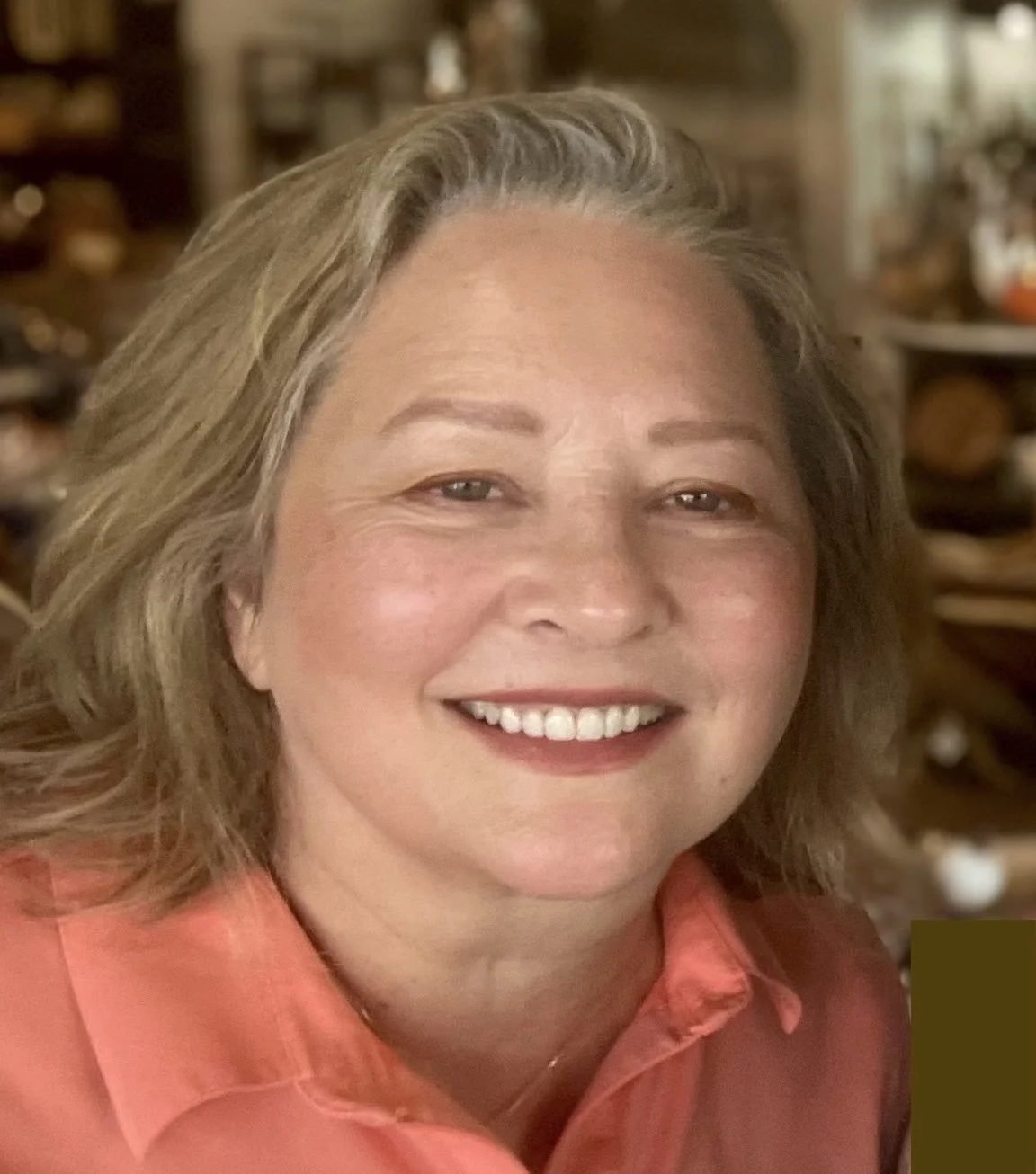 A woman with blonde hair smiling, wearing a coral-colored top, in a warmly lit indoor setting.