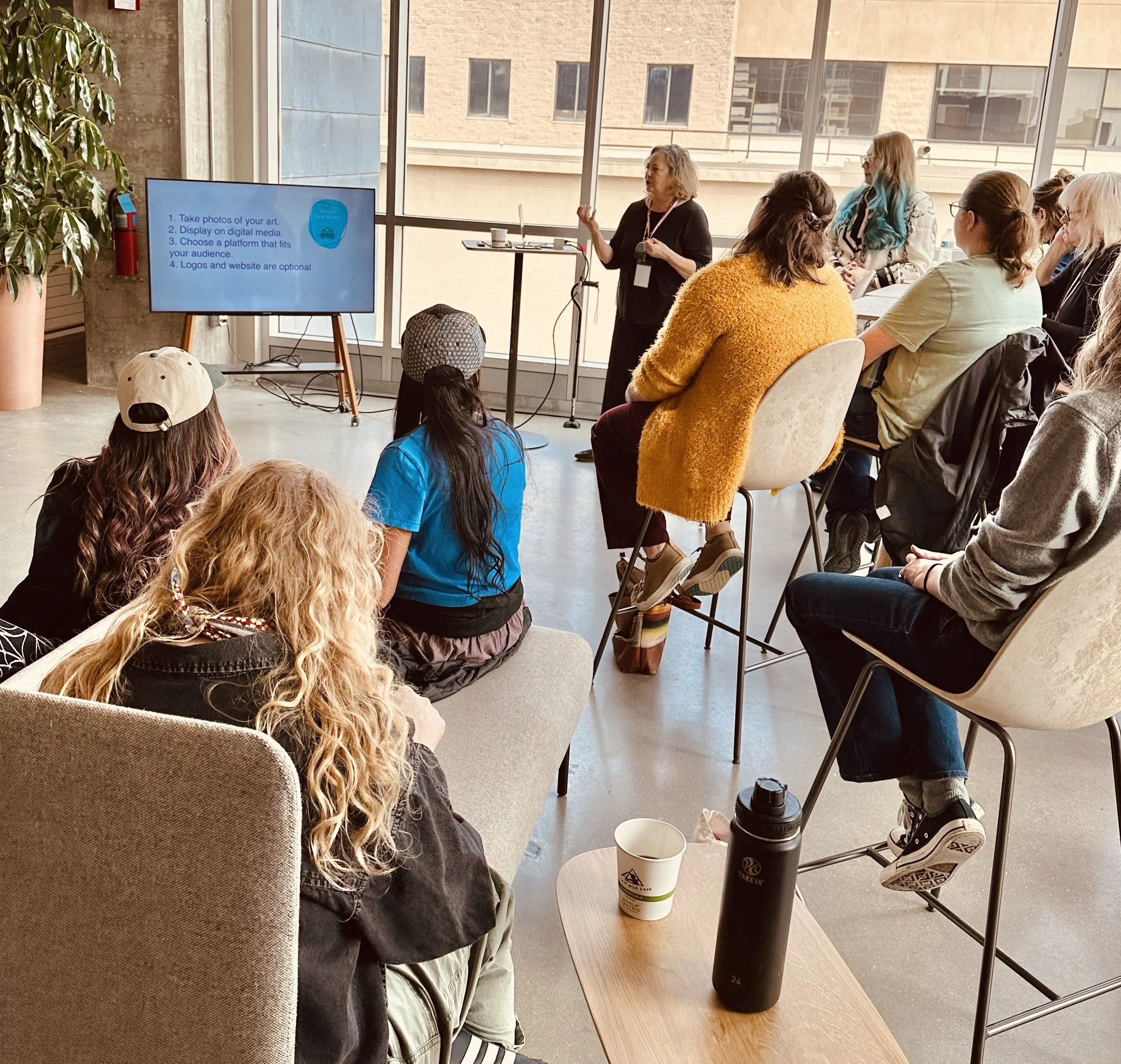 Woman giving presentation to a group of people in a bright room with large windows; on the left, a large screen displays a slide with instructions about sharing art online.