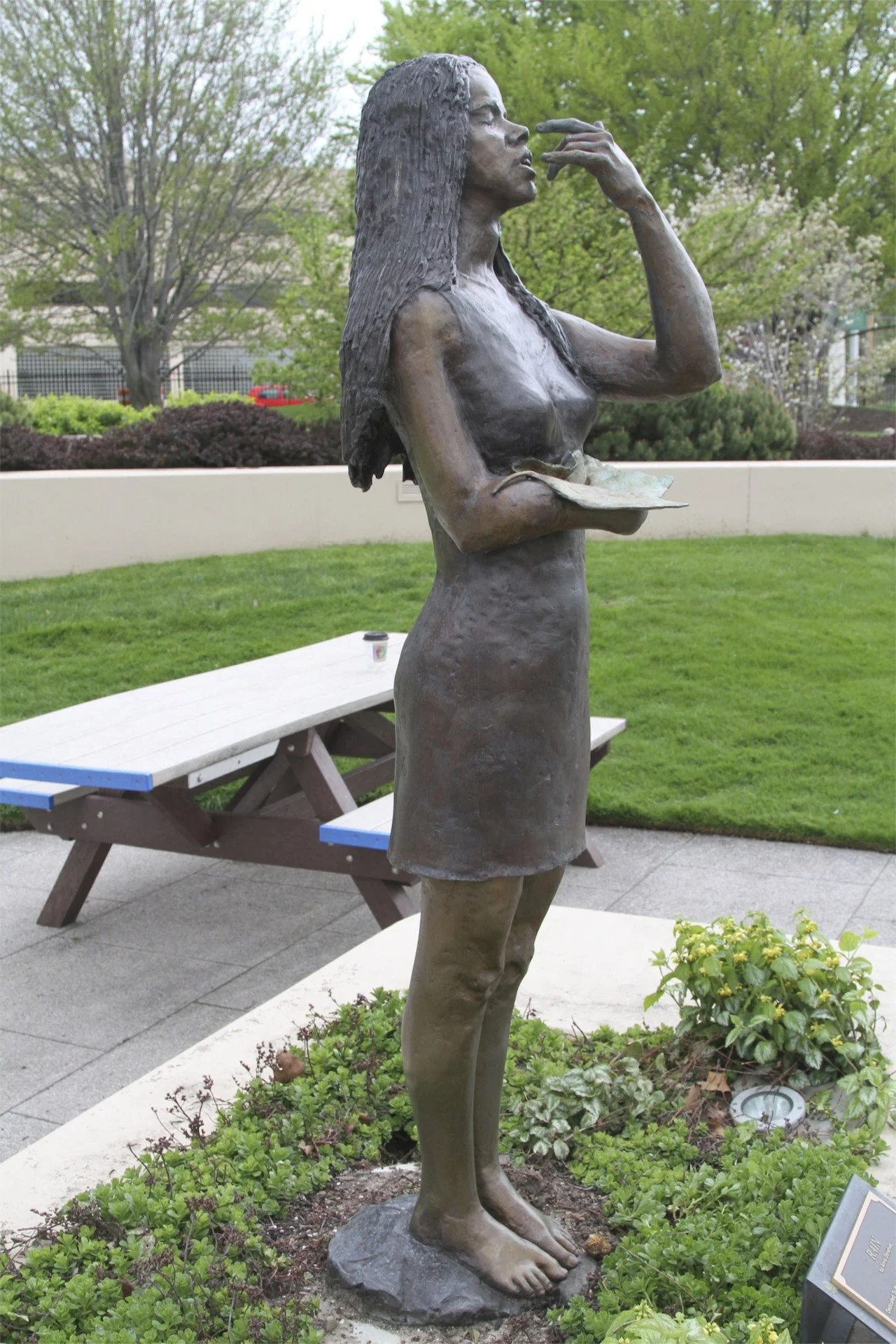 Bronze statue of a woman with long hair, standing outdoors on a rock, with one hand touching her face and the other holding a book. Green trees and a bench are visible in the background.