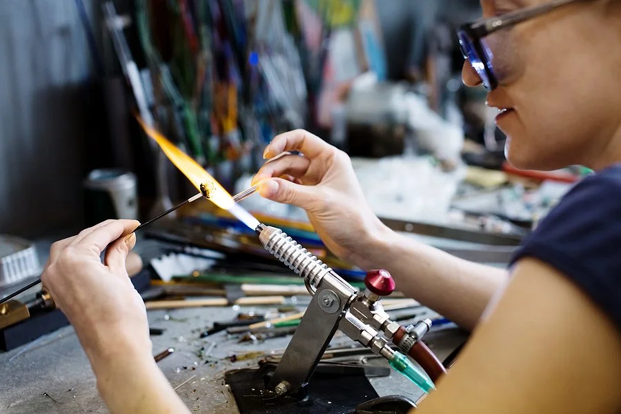 A woman working on glass blowing, holding a glass piece over a flame at a workshop table filled with tools and supplies.