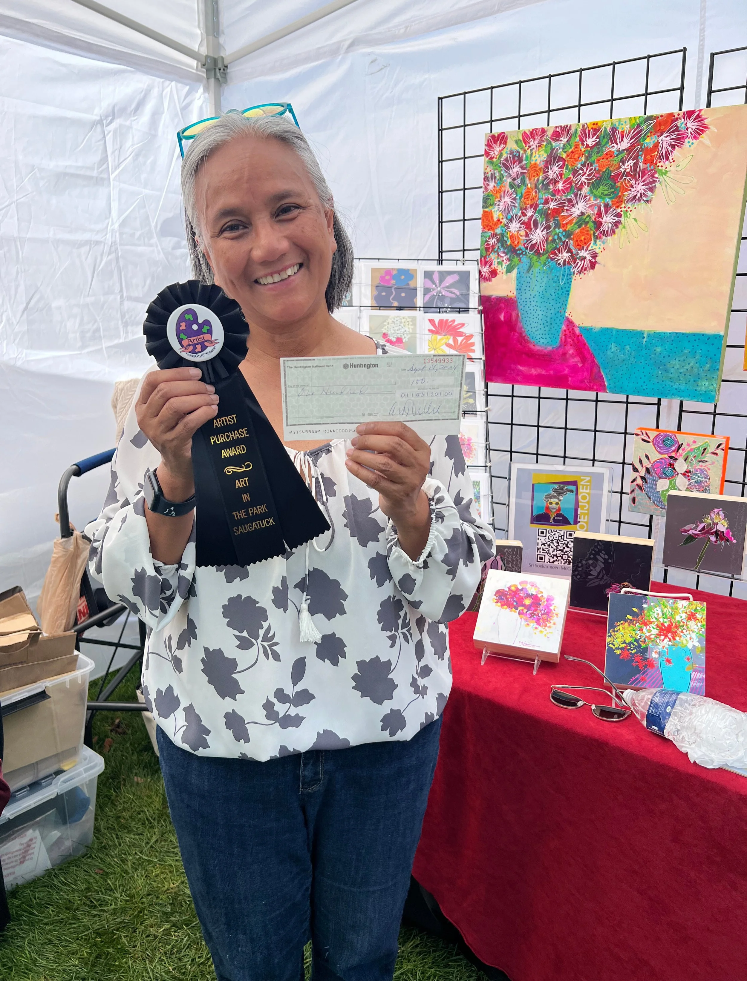 Woman smiling at art fair holding a black award ribbon with a colorful logo and text, standing in front of framed colorful artwork including a large painting of a blue vase with flowers, and smaller art pieces displayed on a red tablecloth, inside a white tent with grass visible on the ground.