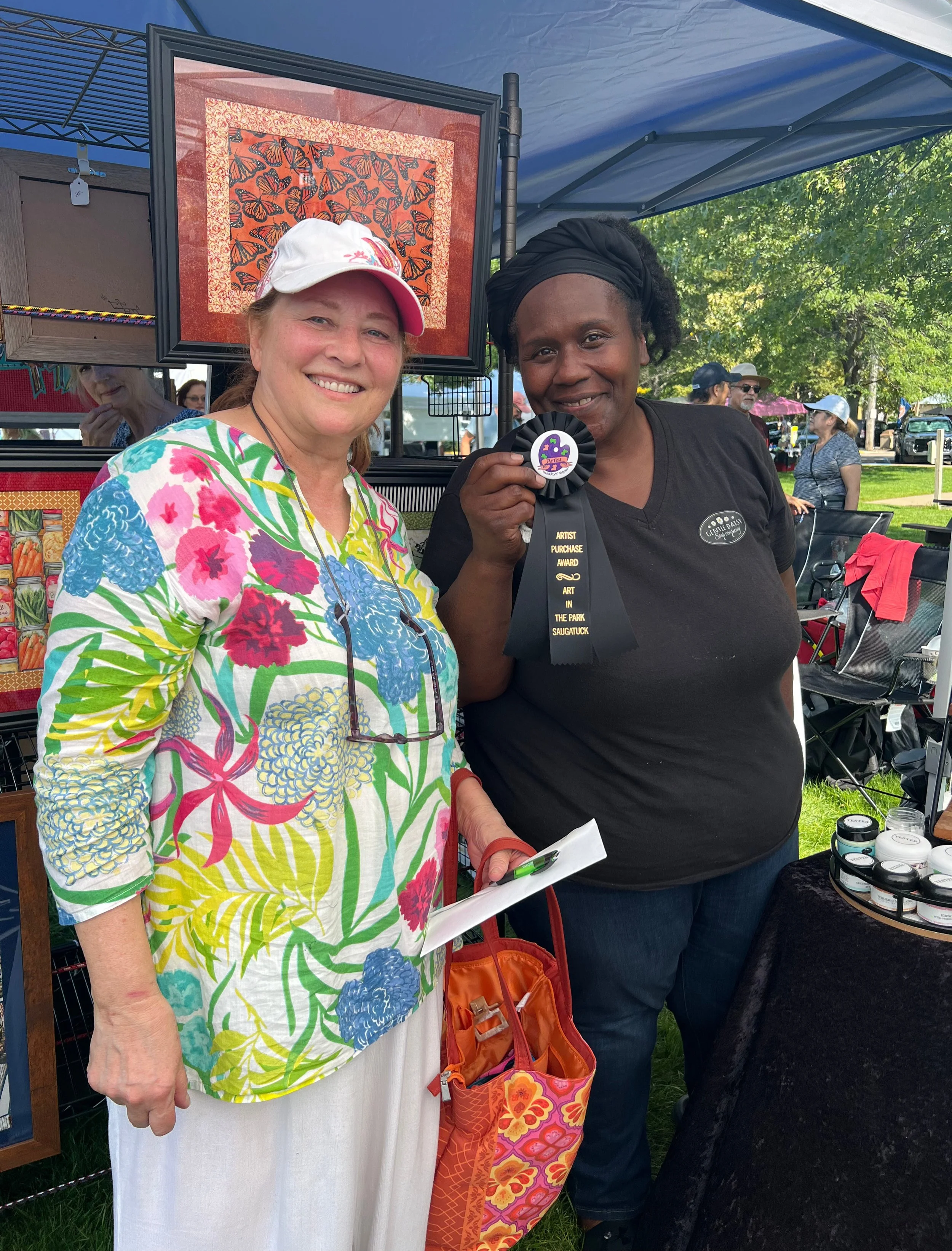 Two women stand outdoors under a blue canopy, smiling at the camera. One woman wears a floral blouse and a white hat, holding a notebook, and the other woman, with a black headwrap, holds a ribbon award. Behind them is a framed piece of artwork with butterflies and other people walking and browsing booths at an event with trees and cars in the background.