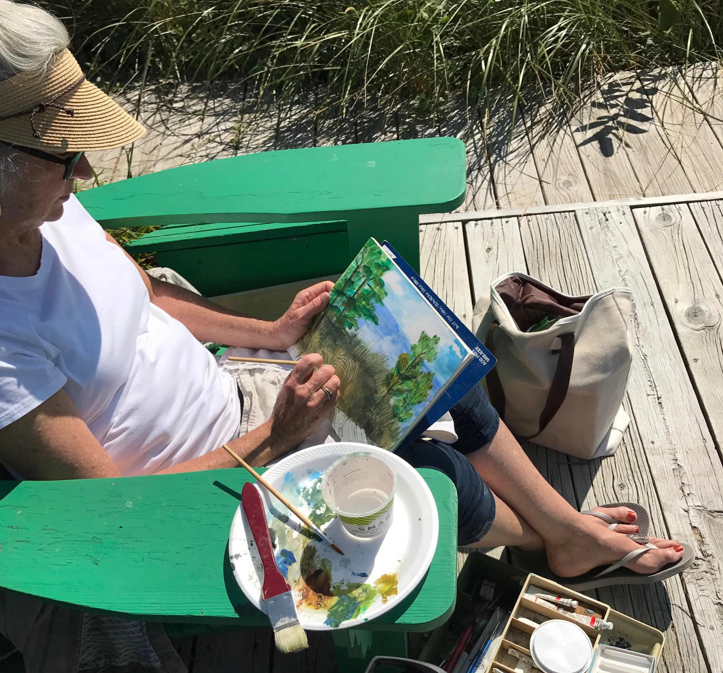 A woman in a straw hat and white shirt is sitting outdoors at a wooden deck, reading a colorful book with illustrations of trees and a sky. She has a paintbrush in her hand, and an open paint palette with green, yellow, and brown paints rests on a green bench next to her. A bucket with painting supplies and a beige bag are nearby.