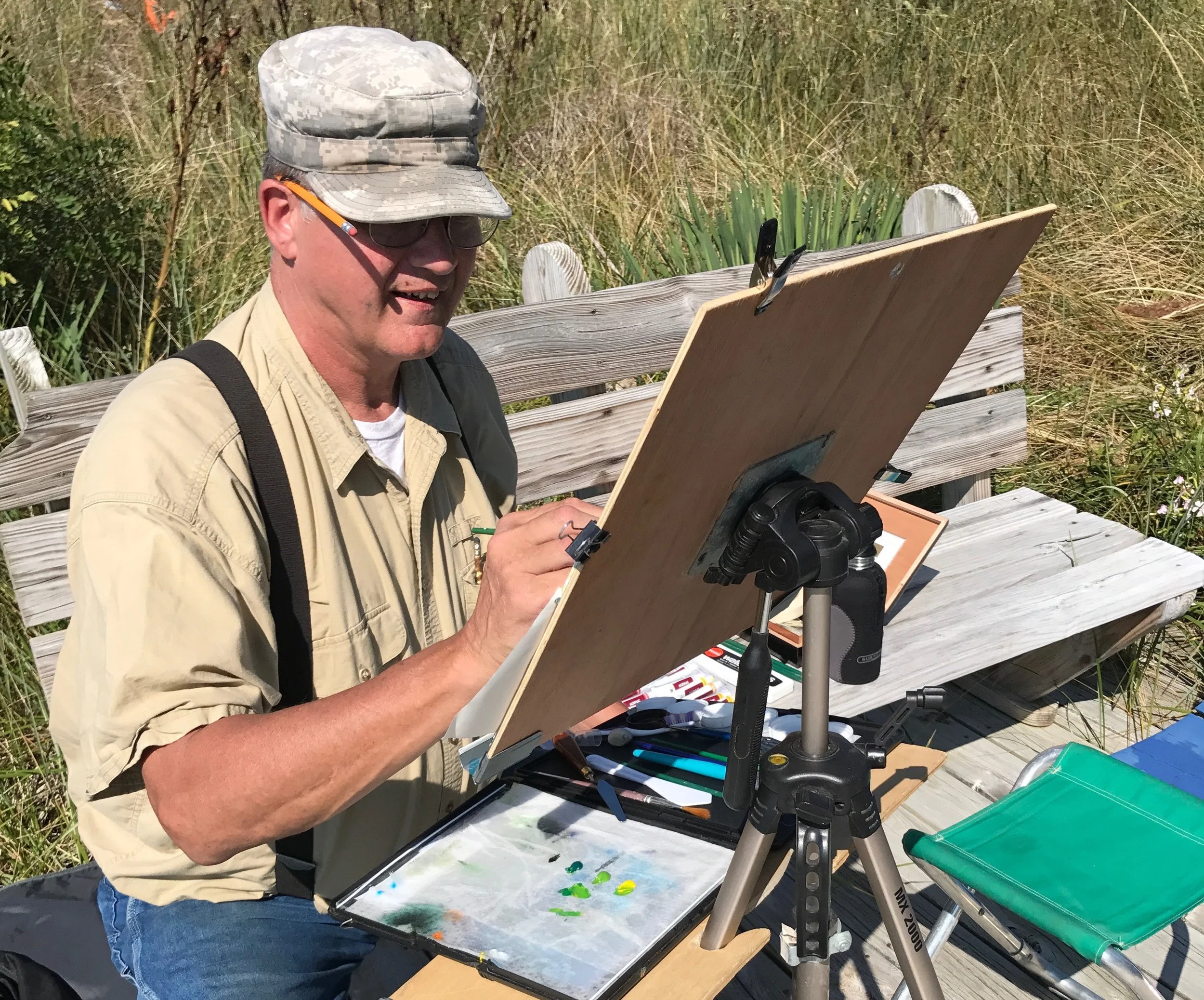 A man in a tan shirt, digital camouflage cap, and sunglasses is painting outdoors on an easel, with a weathered wooden bench and grassy vegetation in the background.
