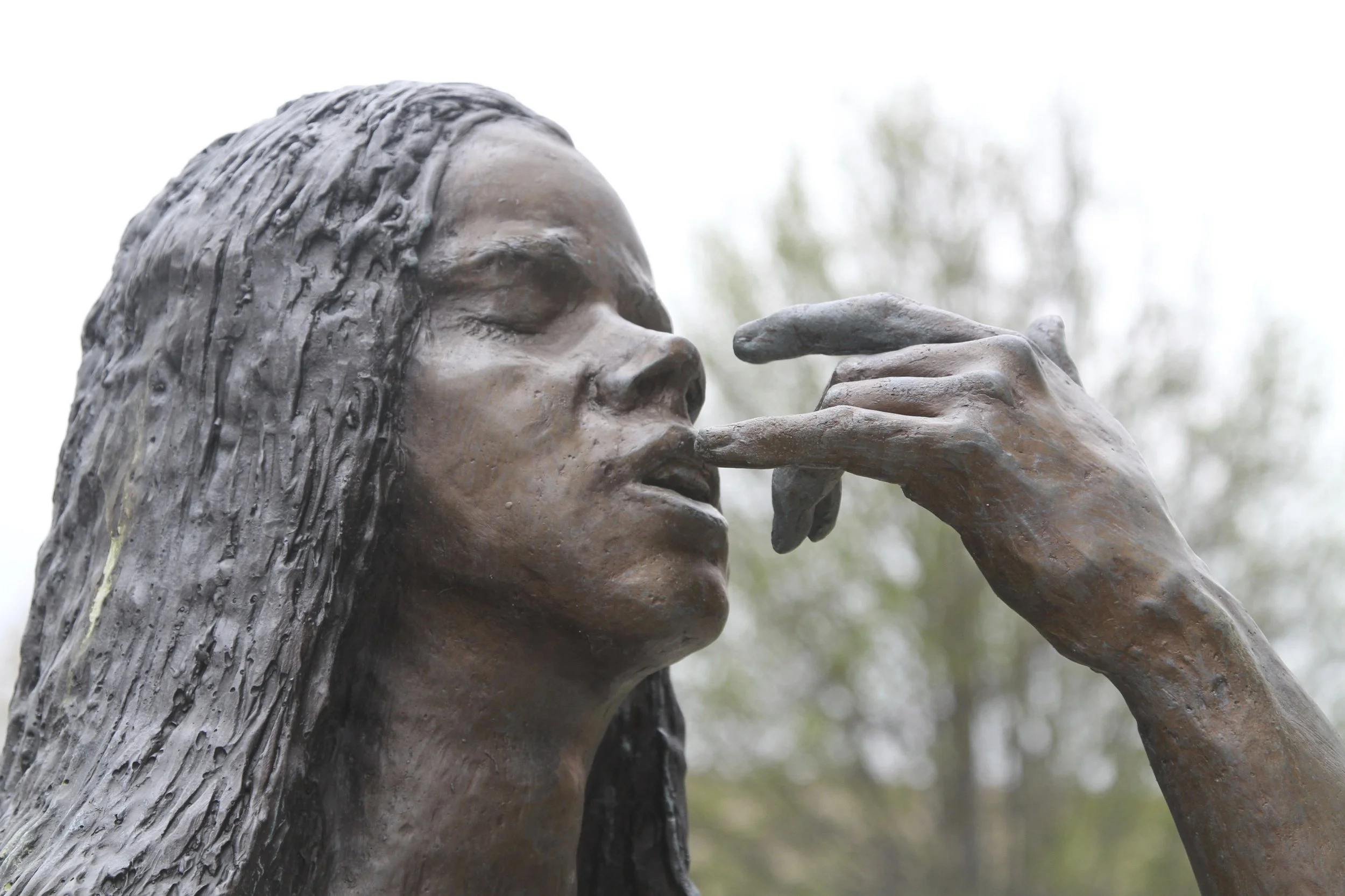 Close-up view of a bronze statue of a woman with long hair, holding her hand near her face, outdoors with blurry trees in background.