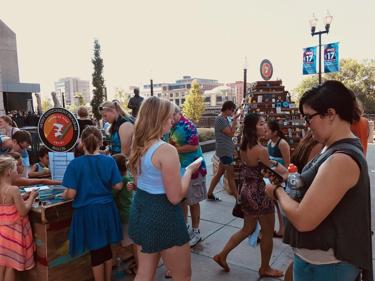 People gathered at an outdoor event with a logo sign, reading 'Artist Enrichment Program, Artist, #66126', and a pyramid of books or items on display, with city buildings and trees in the background.