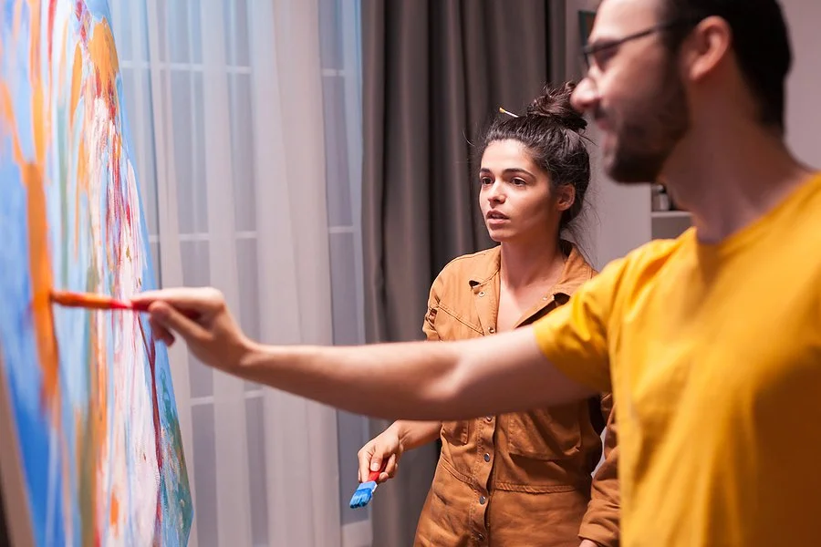A man and woman painting a large colorful mural on a wall in a room with gray curtains and white walls.