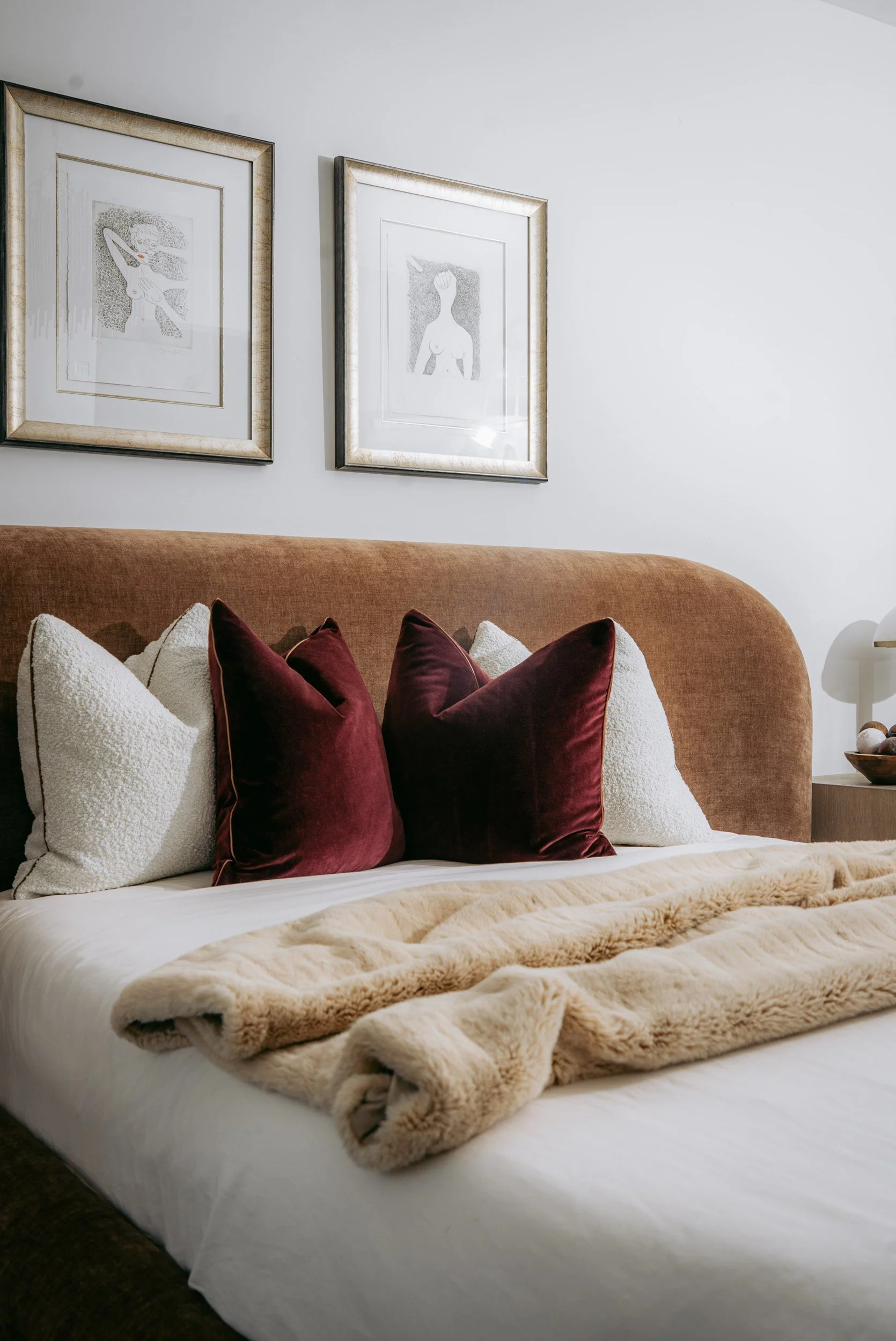 A neatly made bed with white sheets, a beige blanket, and a variety of pillows including white and maroon ones. Behind the bed, two framed artworks hang on a white wall. One artwork depicts a woman with hair covering her face, and the other shows a woman with arms raised. The headboard is upholstered in a soft, salmon-colored fabric.