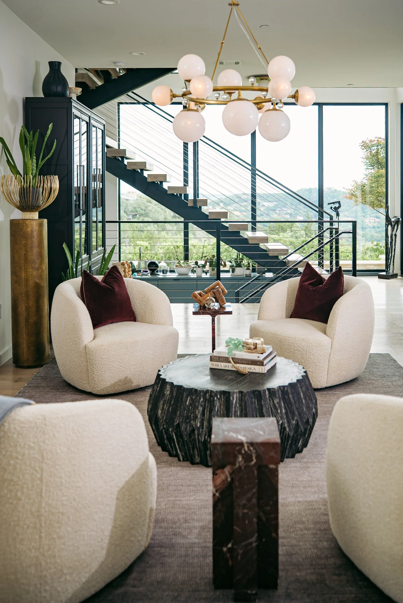 Modern living room with two cream-colored armchairs with maroon pillows, a large black and brown marble side table, and a staircase with black beams and white steps in the background, large windows, a chandelier with multiple white globes, potted plants, and decorative items.