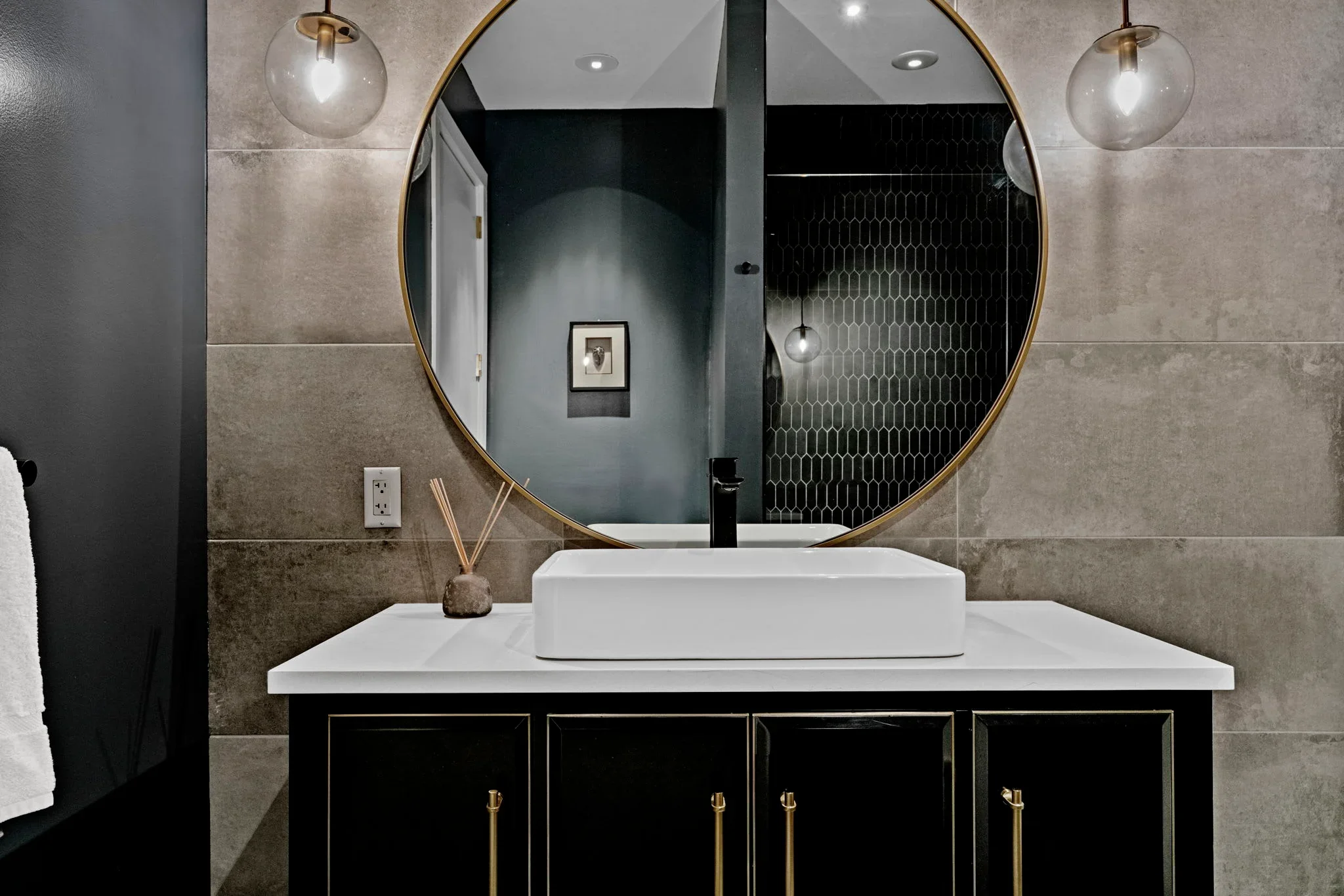 A bathroom with a large circular mirror above a black vanity with gold hardware. A white vessel sink is on the vanity, with a black faucet. There are two pendant lights with clear globes and gold fixtures hanging on either side of the mirror. The wall is tiled with large beige tiles, and there is a small framed artwork visible in the reflection of the mirror.