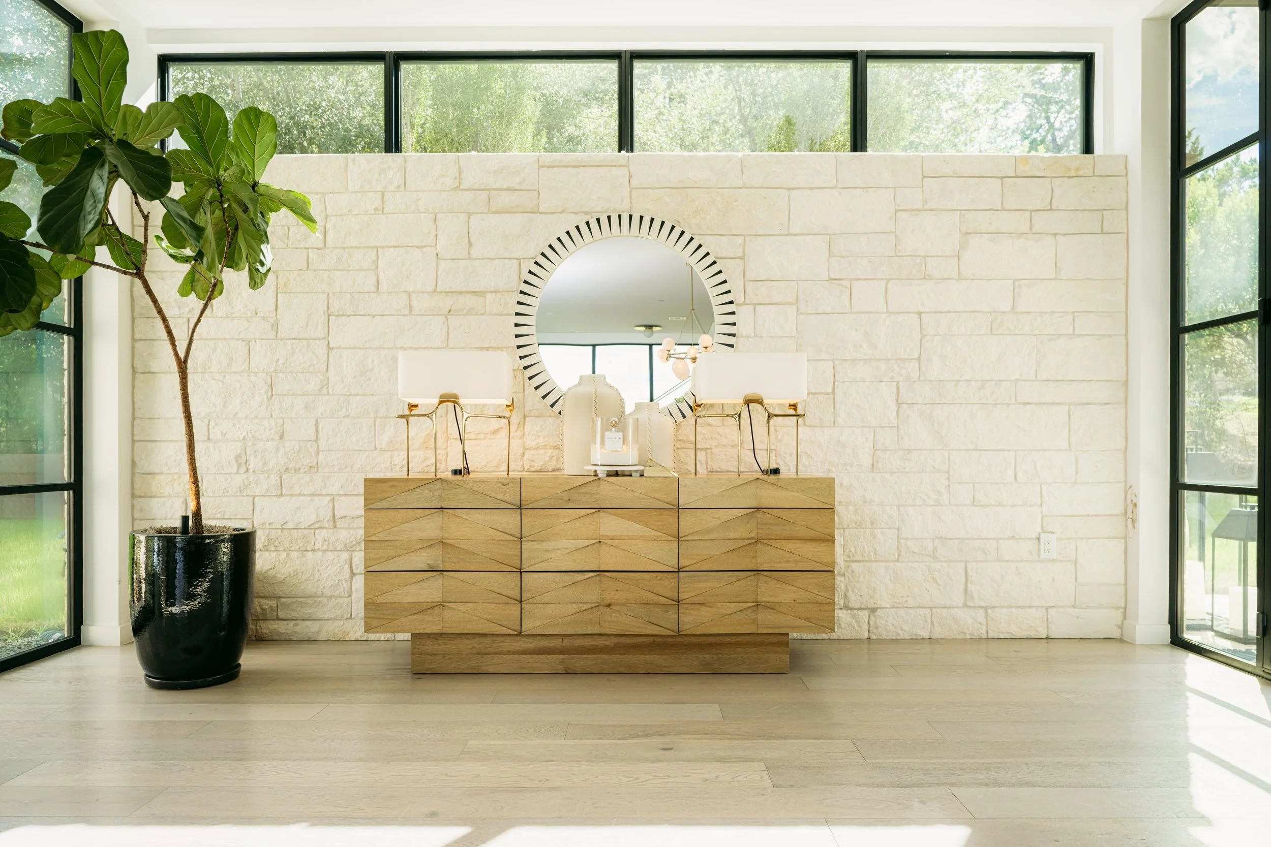 A limestone wall with a carved oak credenza, a sunburst mirror and a fiddle-leaf fig.