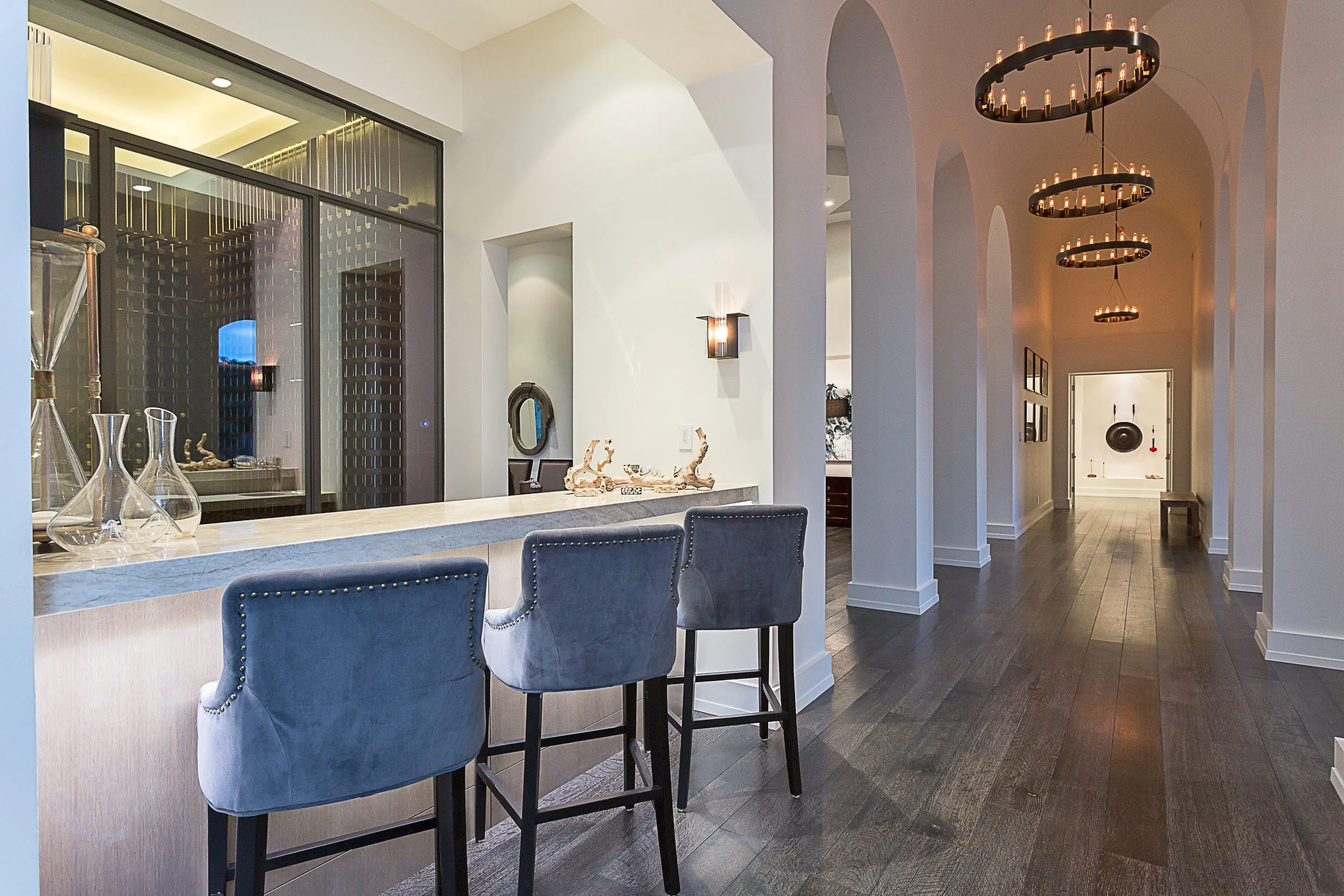 Modern interior with a marble kitchen island, four blue cushioned bar stools, decorative glassware, and a lit chandelier with multiple light bulbs hanging from the ceiling. White walls with modern light fixtures, and a hallway leading to other rooms.