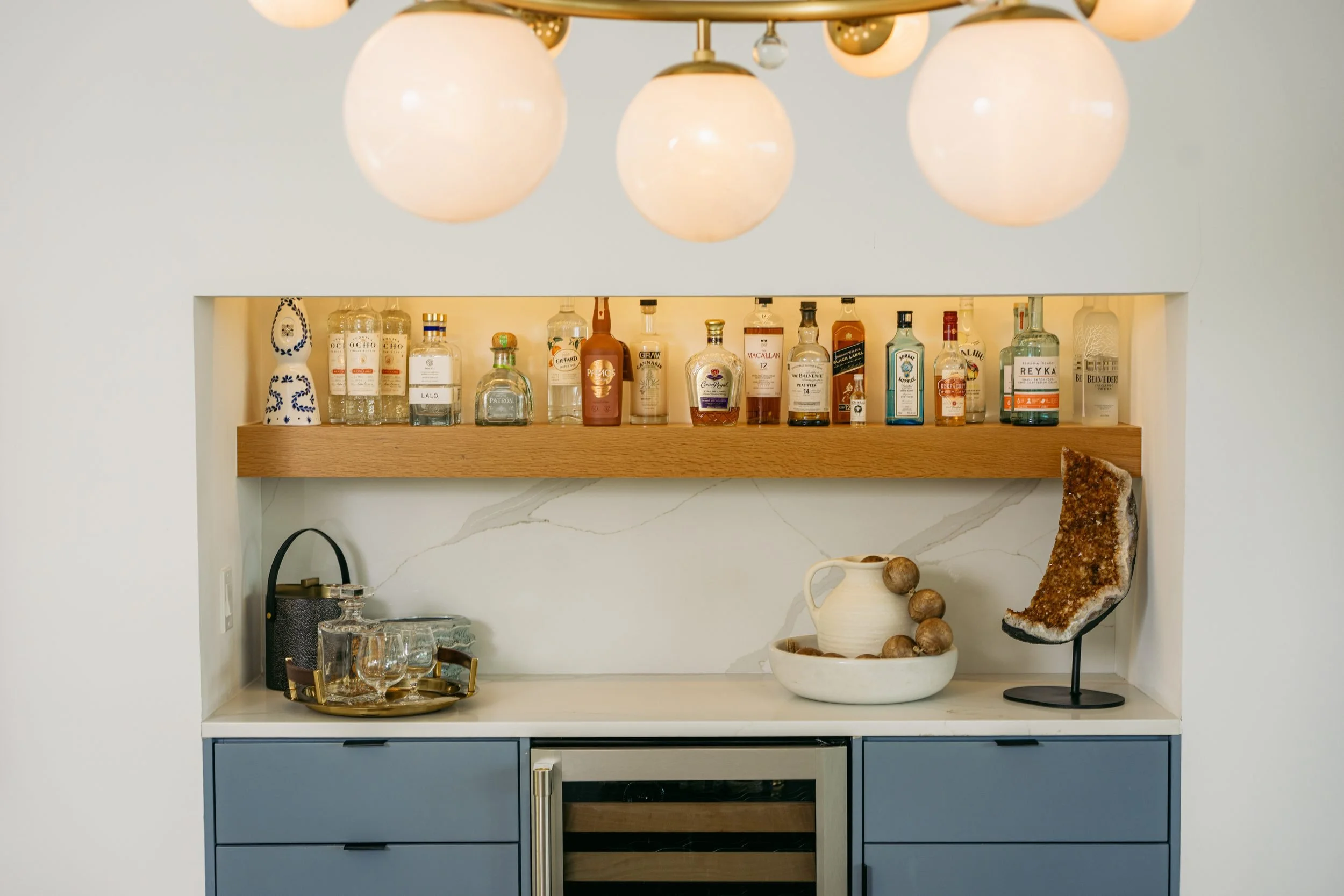 A lit bar niche with an oak shelf, glassware and blue-gray cabinetry.