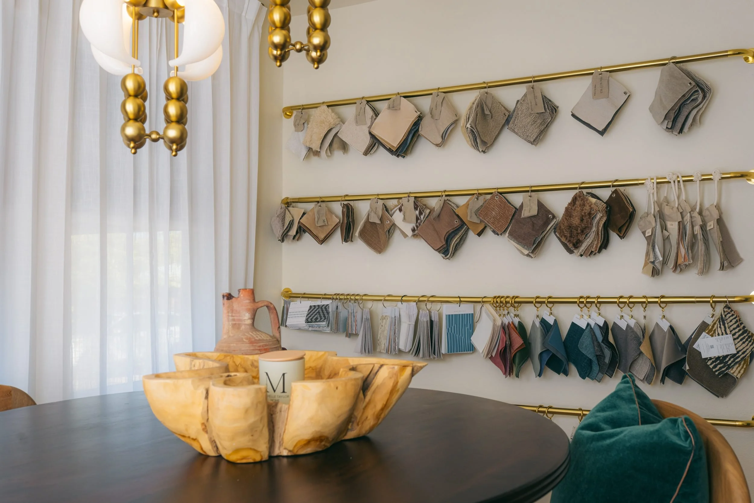 The trade room sample wall — leathers, linens and wallcoverings on brass rails — above a carved wood bowl.