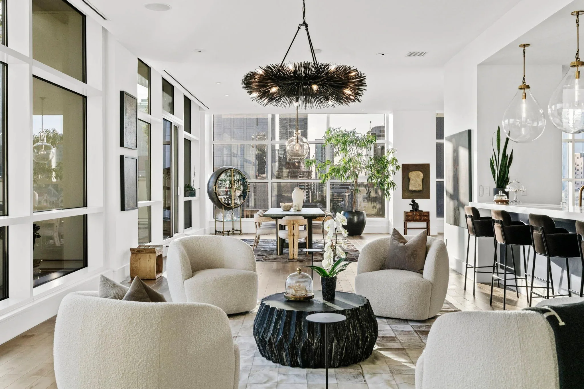 Bright, modern living room with large windows, a black chandelier, and a mix of art, plants, and furniture including a sectional sofa and chairs.