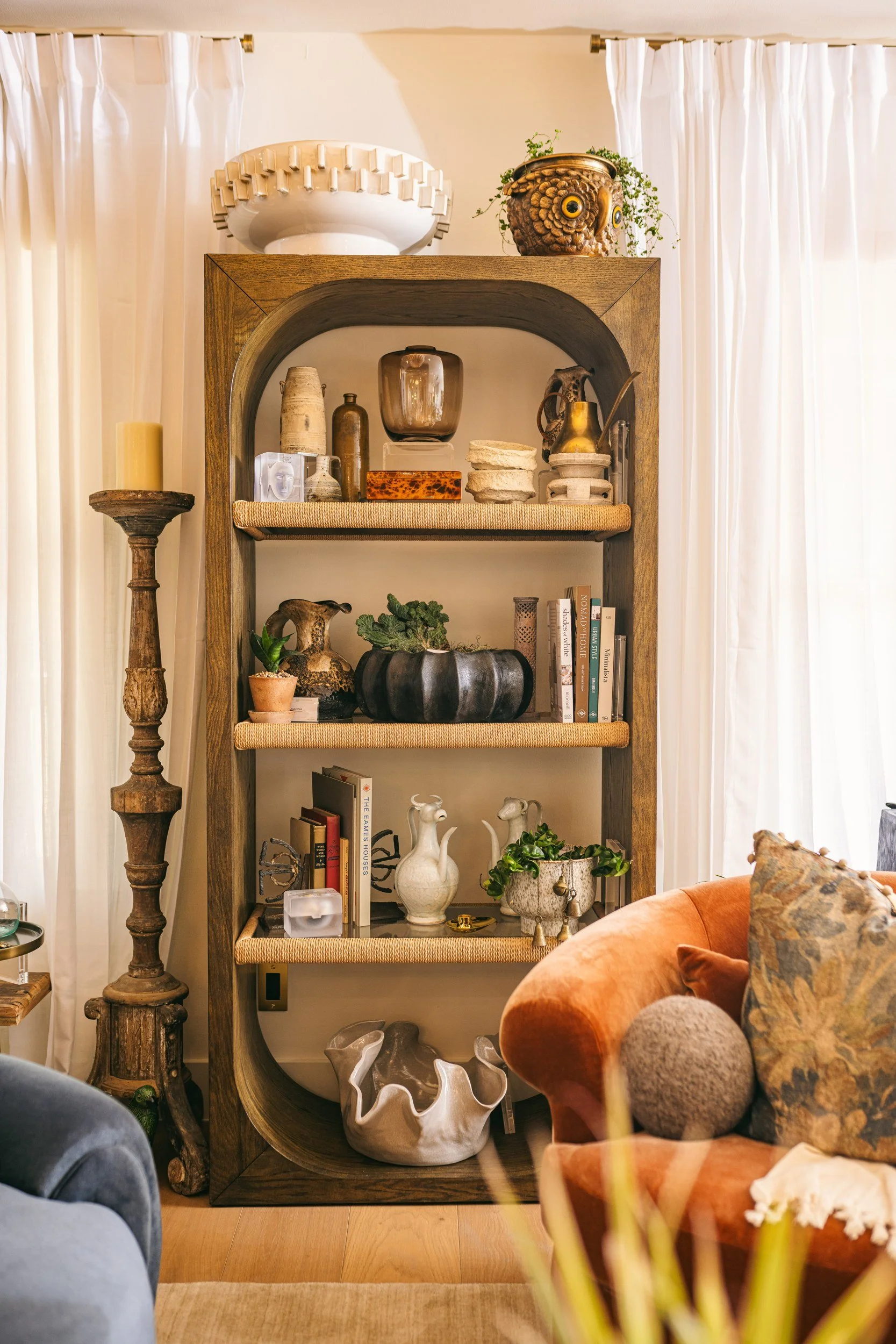 Decorative bookshelf with various ceramics, plants, and books, flanked by a wooden candlestick and a velvet armchair with a floral cushion, in a cozy living room with white curtains.