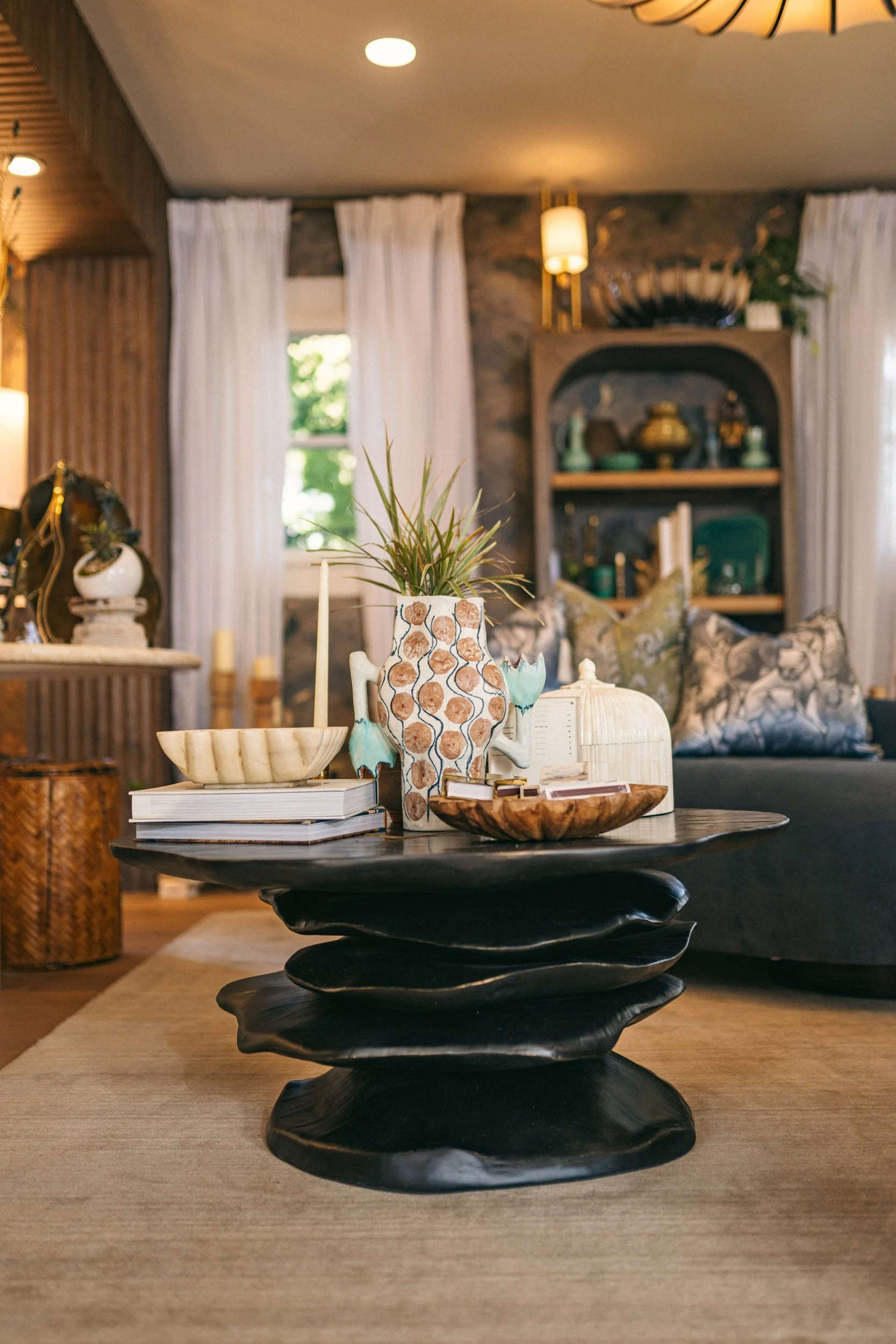 A black wooden tabletop with stacked books and decorative ceramic vases, a beige bowl, and a wooden tray in a warm, inviting living room with curtains, wall art, and interior lighting.