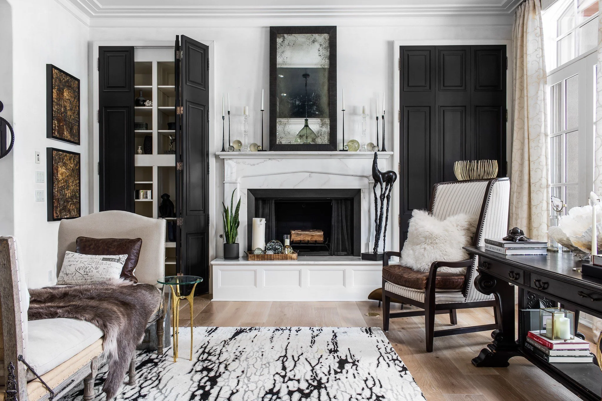 Living room with a white fireplace, black cabinets, a large mirror, and various decorative objects. There are chairs, a patterned rug, and windows with curtains on the right side.