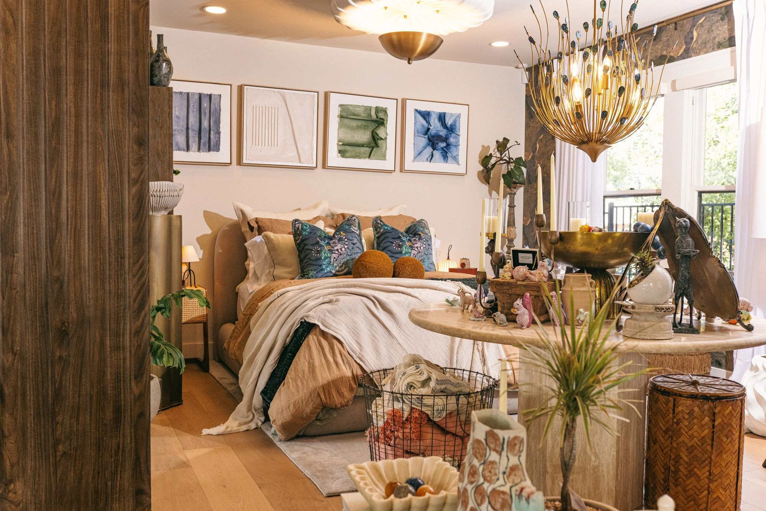 Decorative bedroom with abstract wall art, a bed with patterned pillows, a chandelier, and a variety of decorative objects on a table and nearby surfaces.