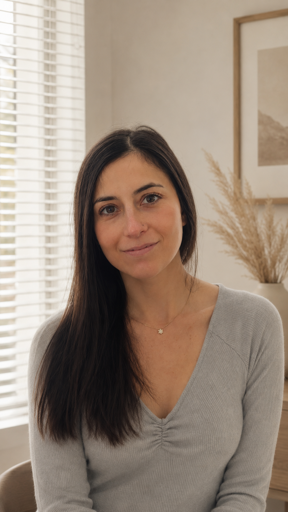 A woman with long, dark hair and a light gray V-neck sweater, smiling softly, sitting in a room with natural light, white blinds, framed artwork, and a beige vase with dried plants in the background.