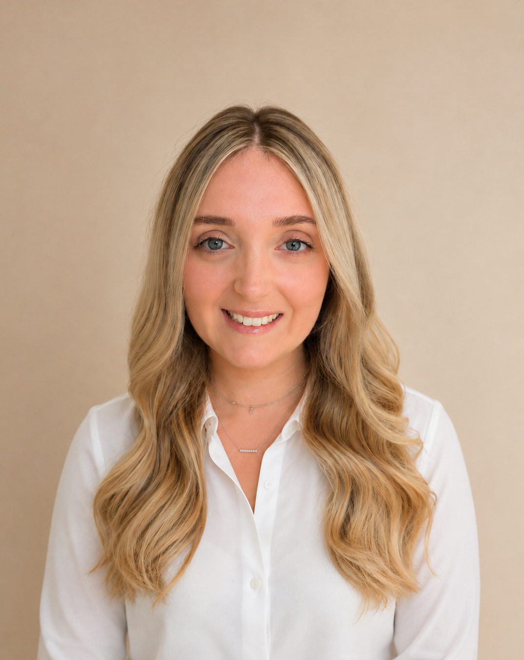A young woman with long blond wavy hair, blue eyes, and fair skin, smiling while wearing a white button-up shirt and layered necklaces, standing against a plain beige background.