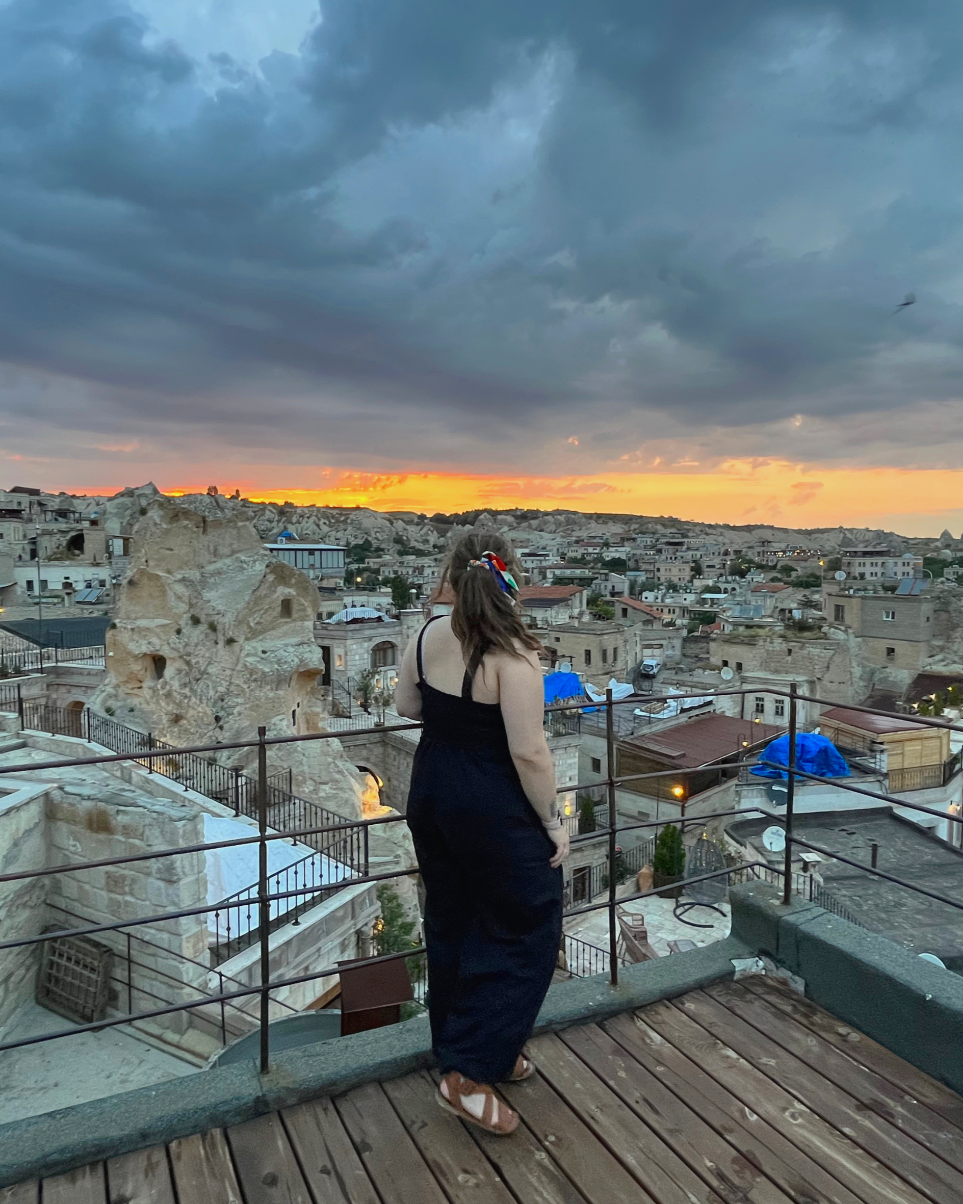 A woman with a bow in her hair looking out over a cityscape at sunset, with cloudy sky and rocky terrain in the background.