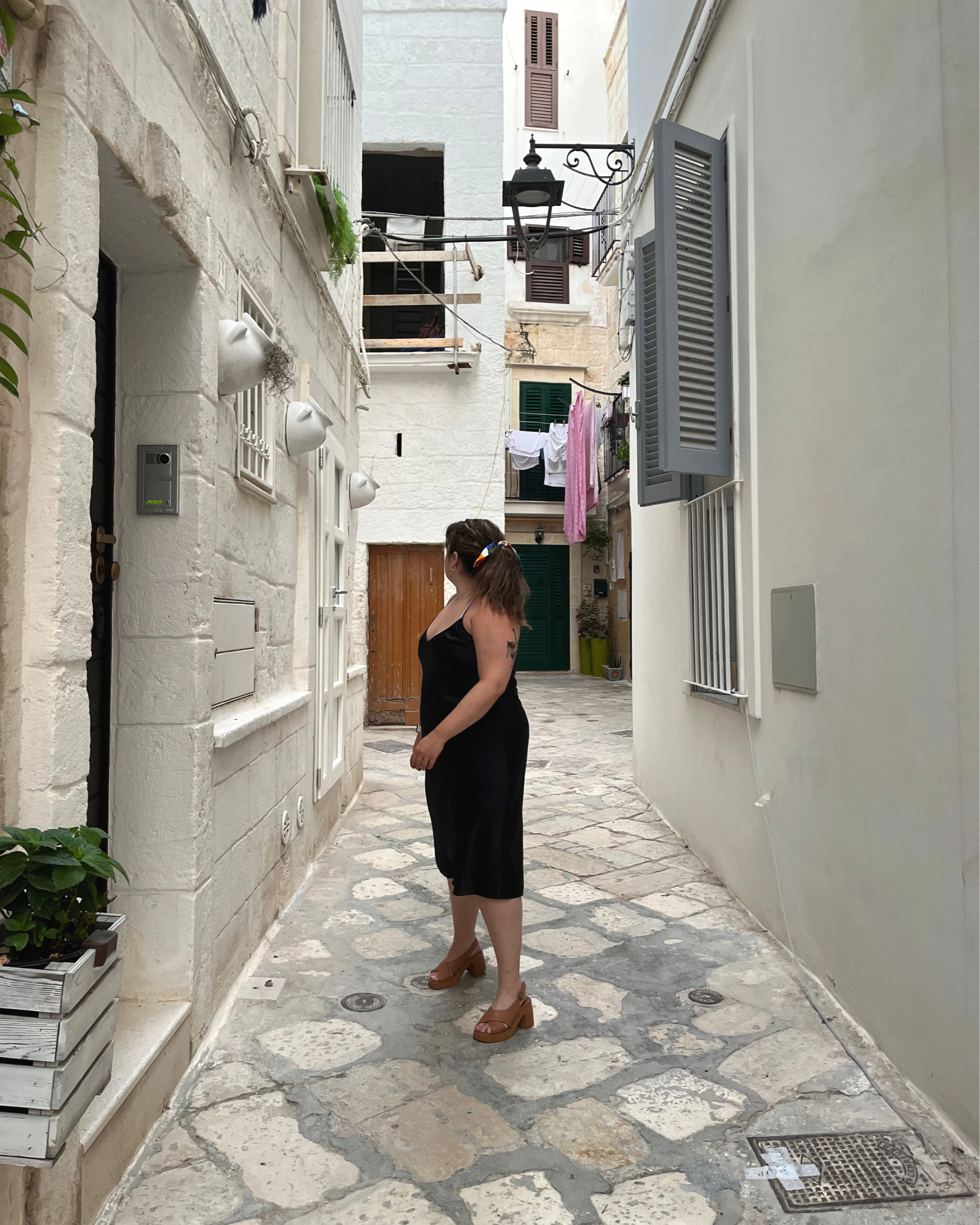 A woman standing on a stone-paved alleyway in a European city, wearing brown platform heels, a black dress, and a colorful headband. The alley is narrow, with white stone buildings on either side, laundry hanging from a balcony, and a street lamp overhead.