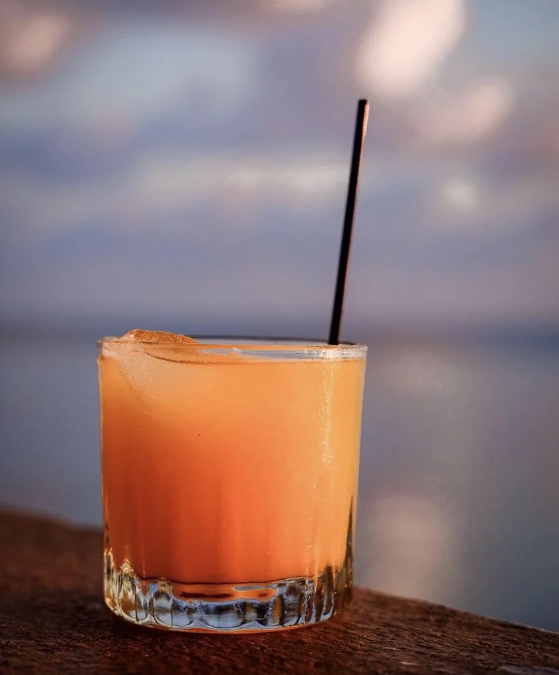 A glass of orange-colored cocktail with ice, garnished with a black straw, on a wooden surface with a blurred ocean and cloudy sky background.