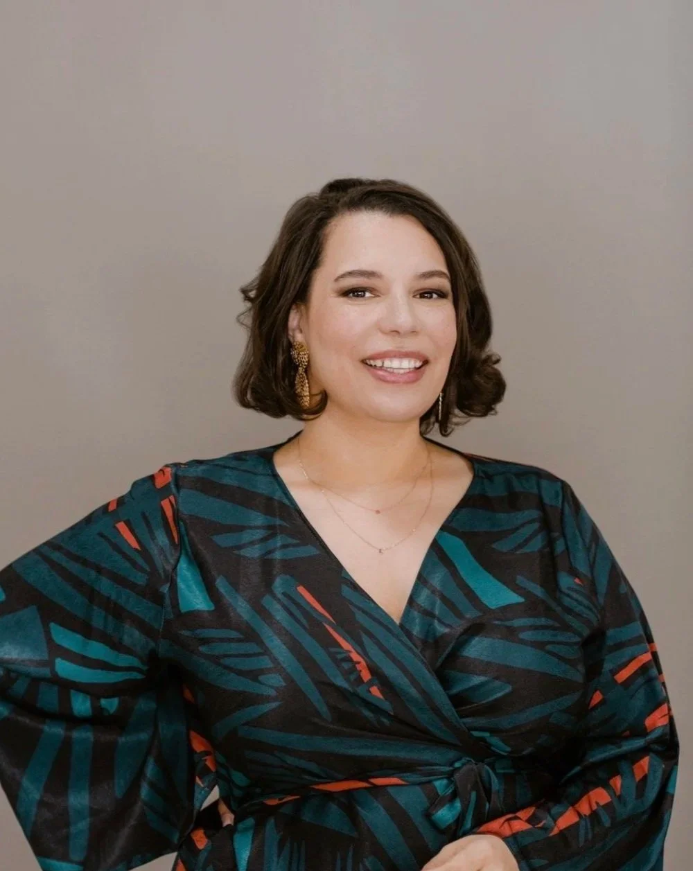 A woman with shoulder-length dark hair, smiling, wearing a dark patterned dress with a V-neck and gold jewelry, standing against a plain gray background.