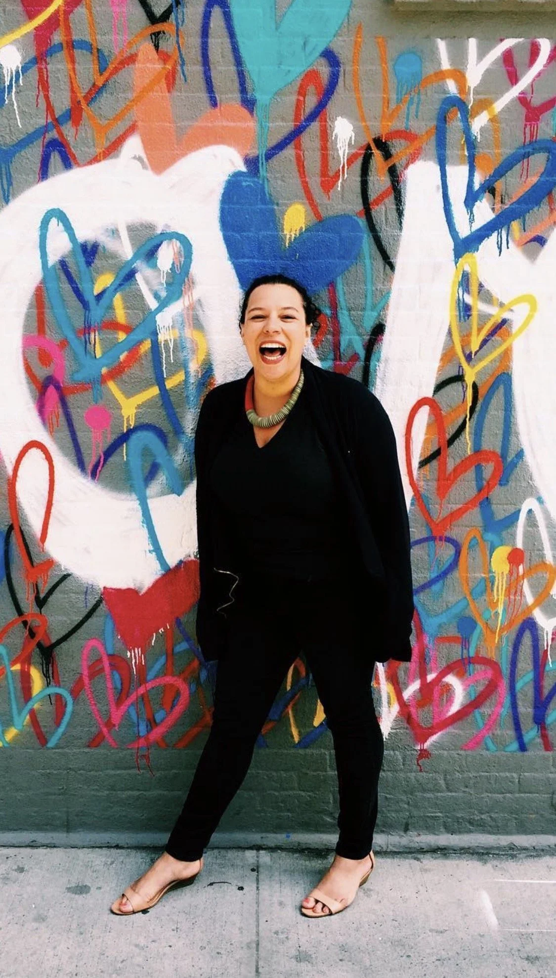 A woman standing in front of a colorful graffiti wall with painted hearts, smiling and laughing, wearing black clothes and beige high heels.