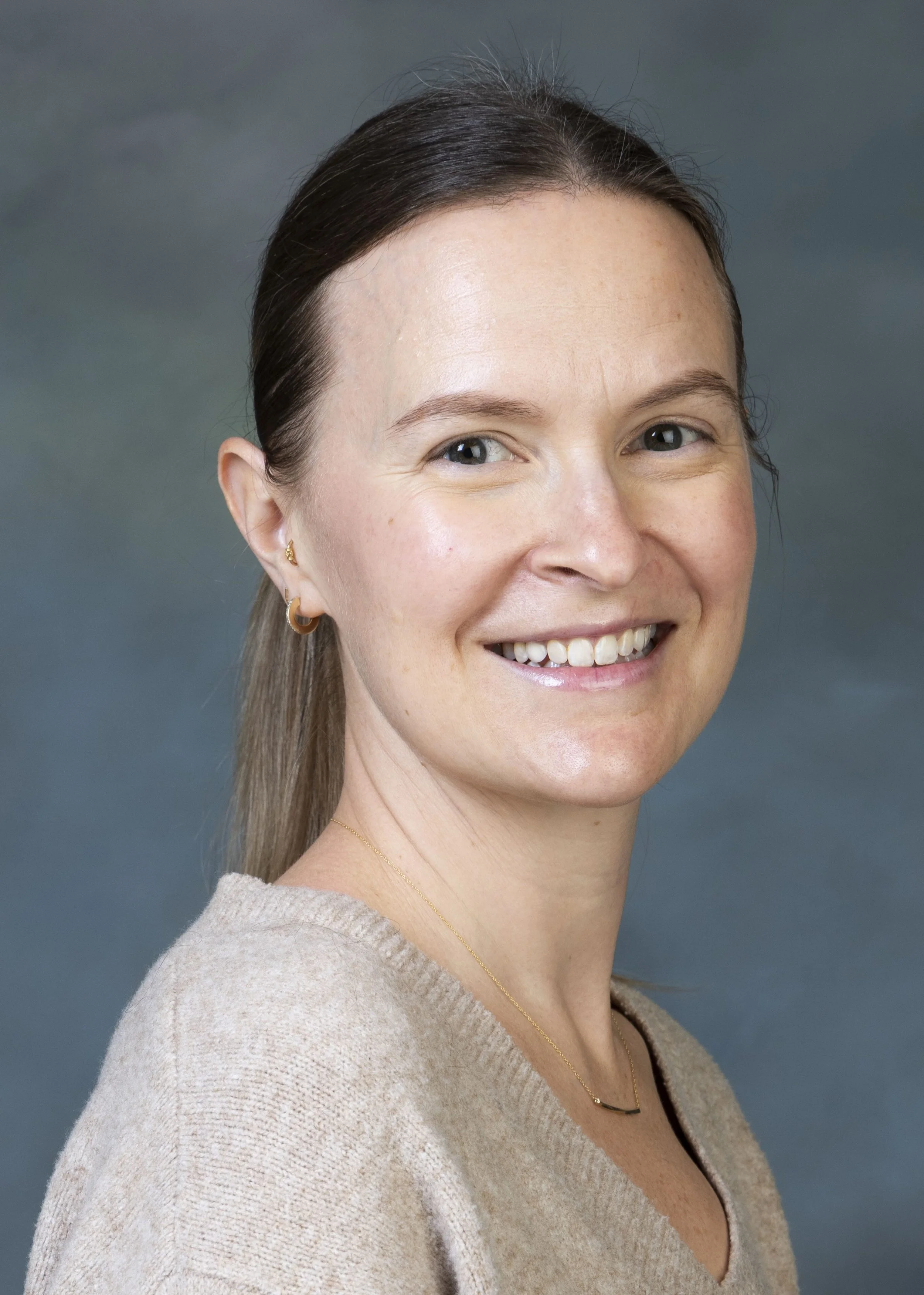 Close-up of a smiling woman with brown hair, blue eyes, wearing gold hoop earrings, a delicate gold necklace, and a beige sweater, against a dark teal background.