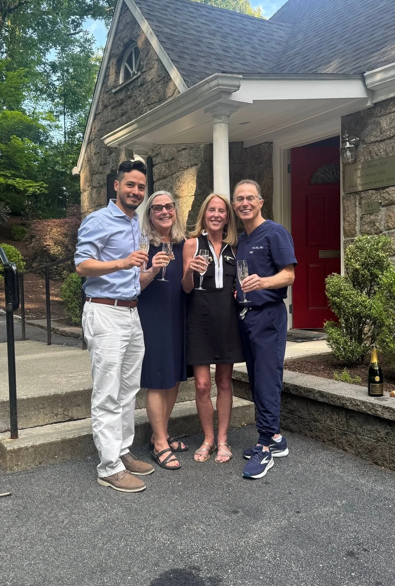 Four smiling people standing together outside a house, holding glasses of champagne, celebrating. The house has a stone exterior, a red door, and a small porch.