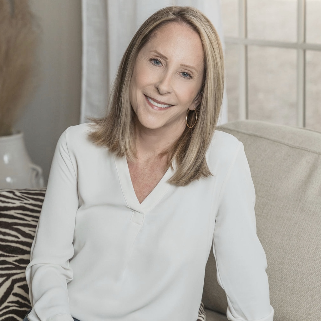 A smiling woman with shoulder-length blonde hair, wearing a white blouse and hoop earrings, sitting on a sofa in a well-lit room with a window behind her.