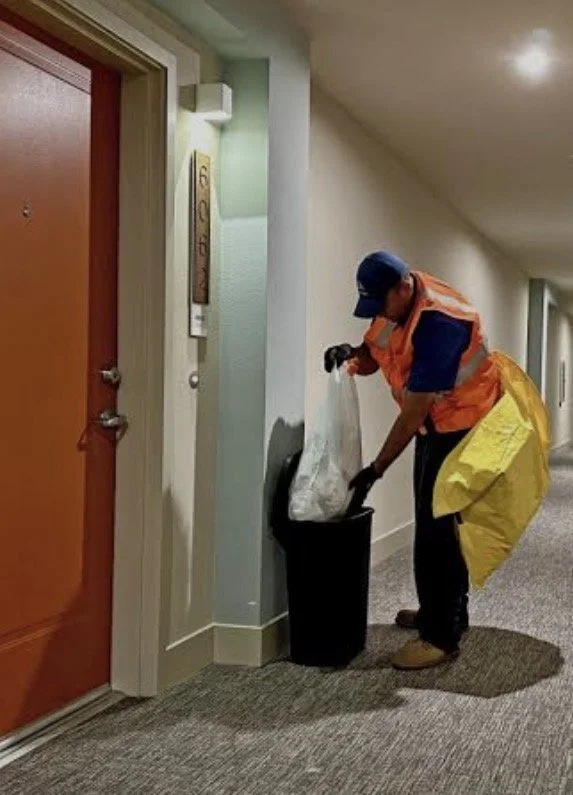 A janitor in an orange and yellow uniform is emptying a trash can in a hallway near an apartment door.
