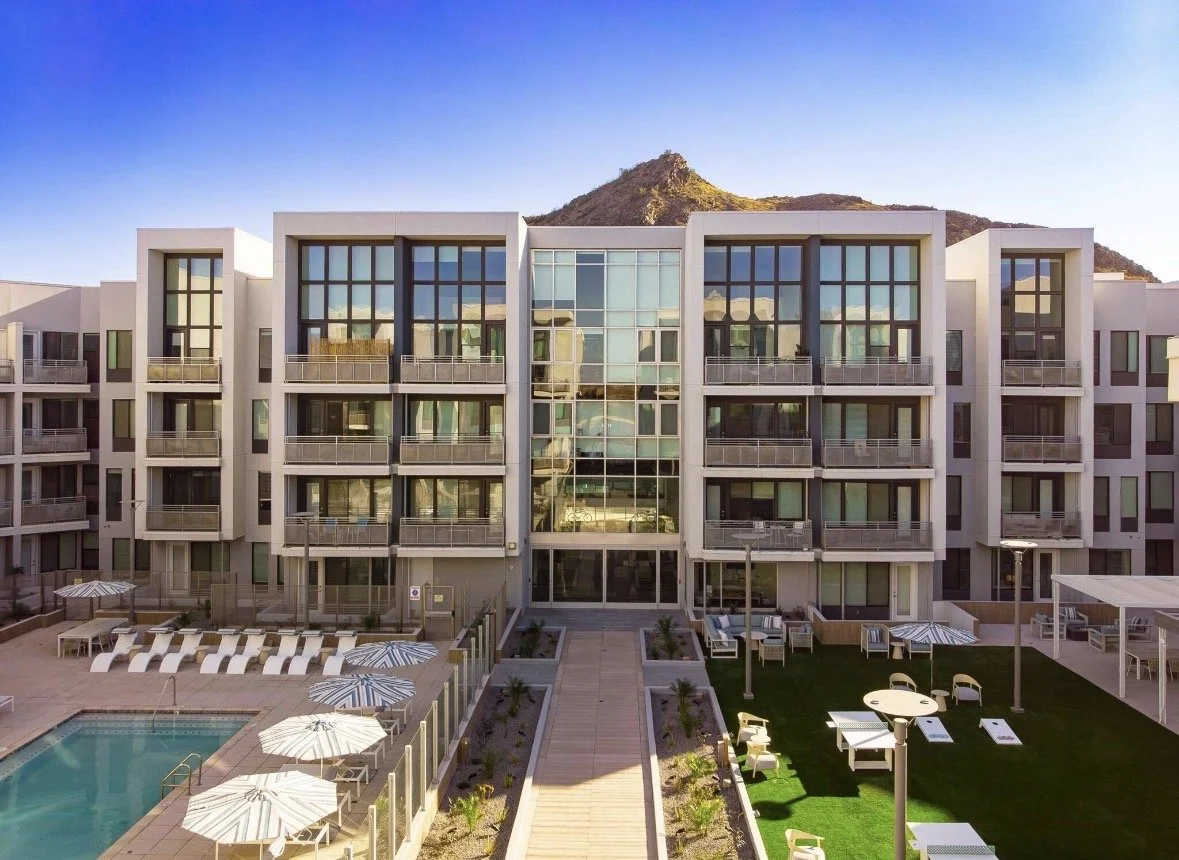 Modern apartment building with a pool and outdoor seating area, mountains in the background.