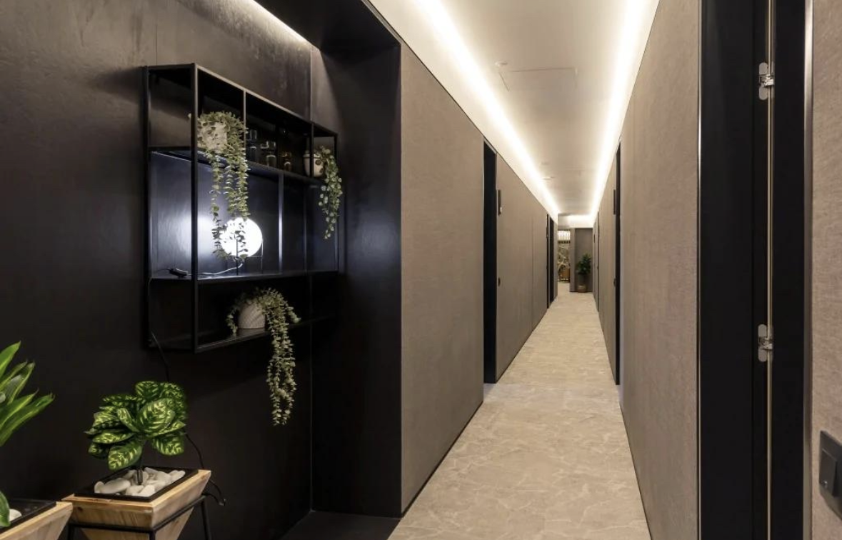 A modern hotel corridor with beige floors, gray walls, and black doors with metallic hinges. There are potted plants and decorative shelving with plants on the left side of the hallway, illuminated by built-in ceiling lighting.