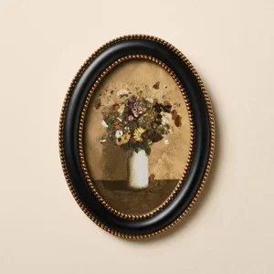 Oval framed painting of a flower bouquet in a white vase on a beige background.