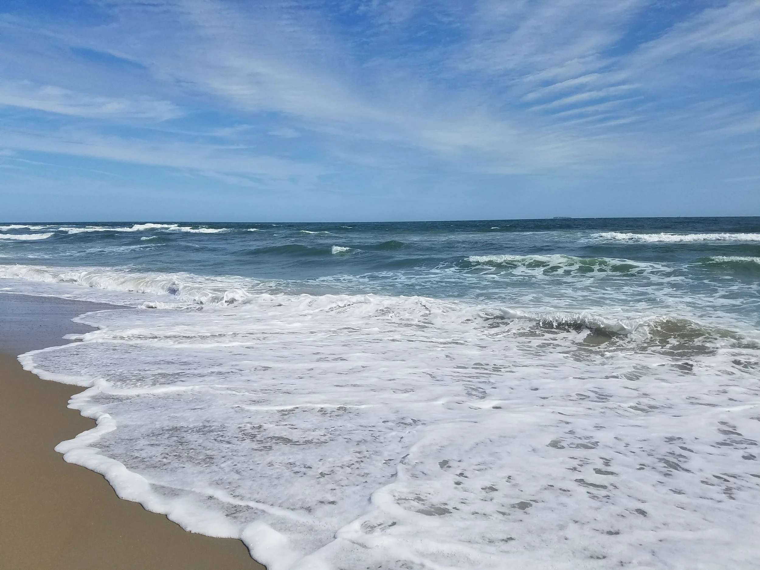 A photo of a wave breaking on a beach. Blue sky overhead, brown sand, and white foam.