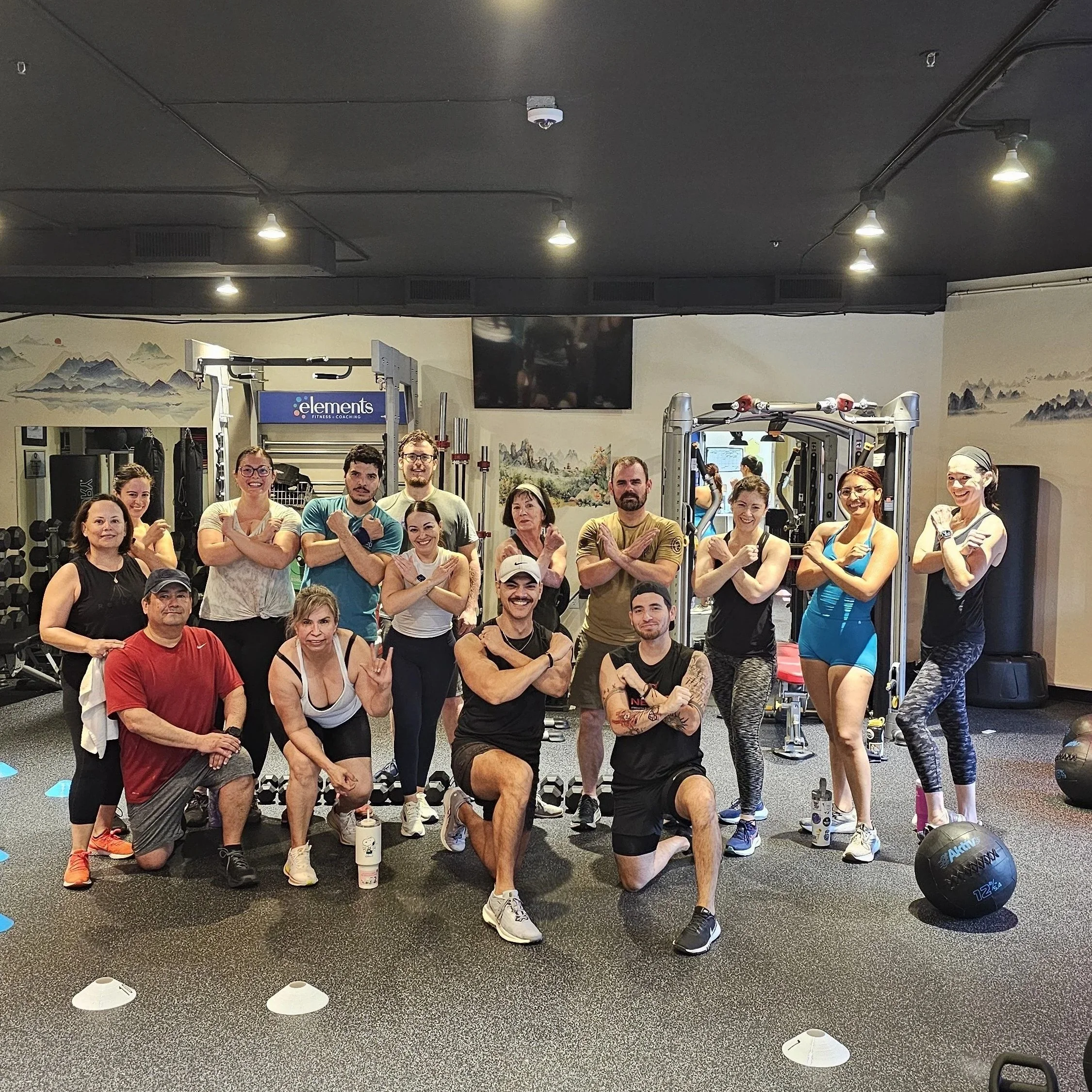 Group of diverse people in a gym posing together with crossed arms, smiling, surrounded by workout equipment and fitness posters.