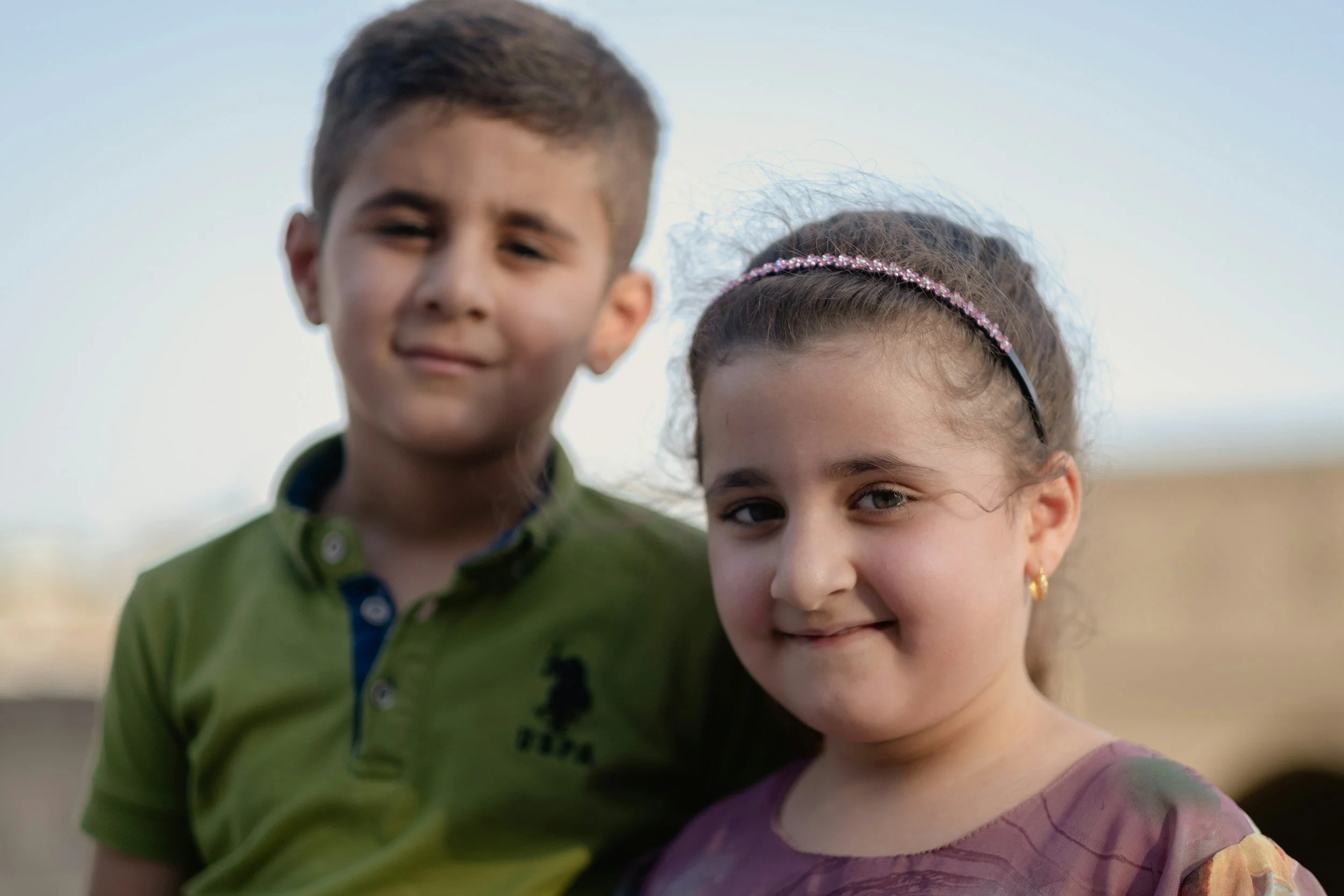 Close-up of a boy and girl outdoors, smiling softly, with the sky in the background.