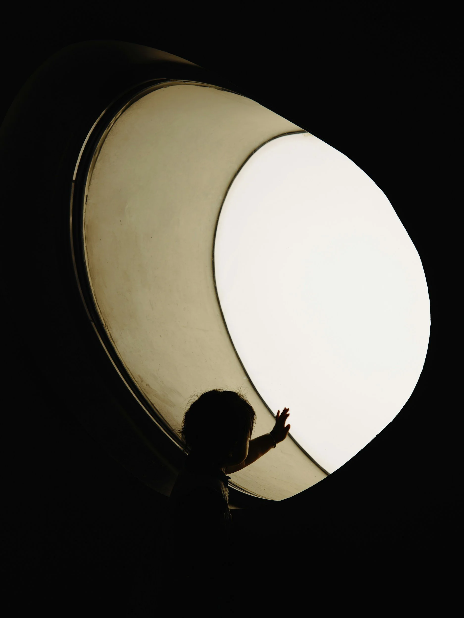 A silhouette of a child reaching out towards a bright, curved light source or window in a dark space.