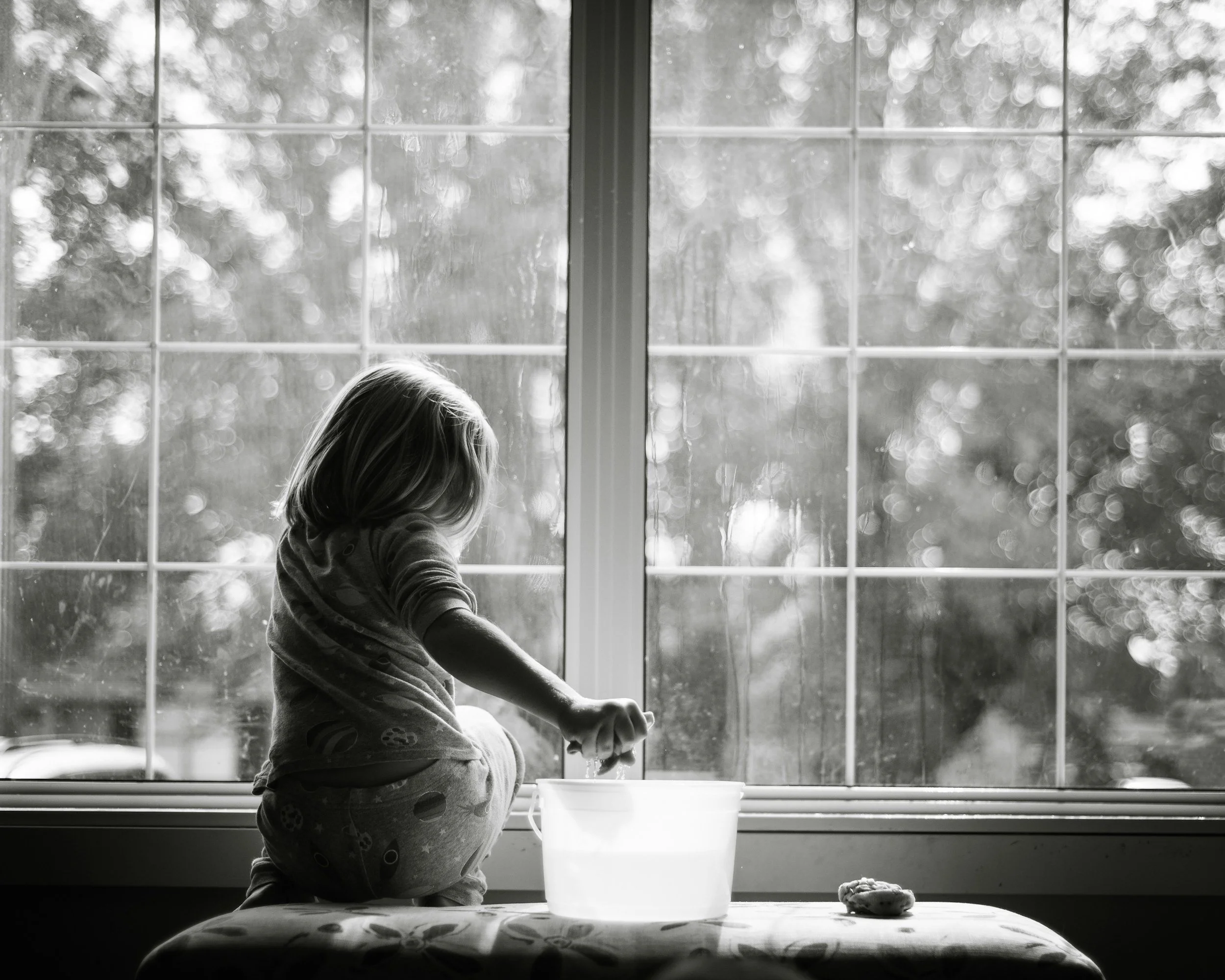 A young girl with long hair, wearing pajamas, sitting at a table by a window, holding a spoon over a bowl, with a plastic container nearby, likely during daytime.