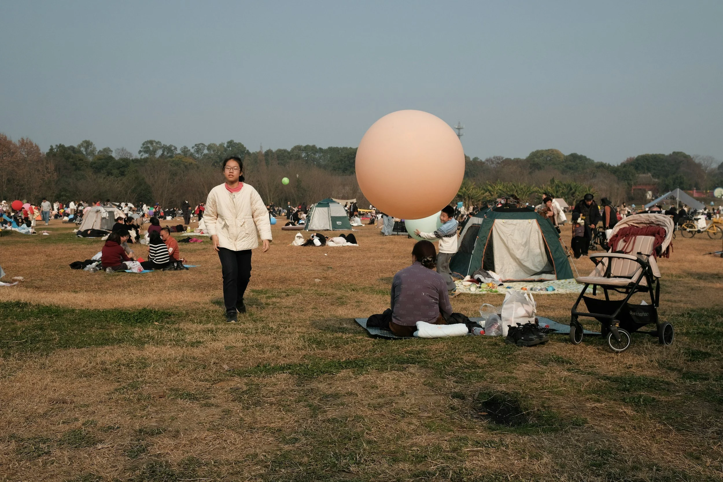 A large open field with numerous people gathered, some sitting on blankets and some walking. Several tents and balloons are visible, and a boy is holding a big balloon while walking. The sky is clear.
