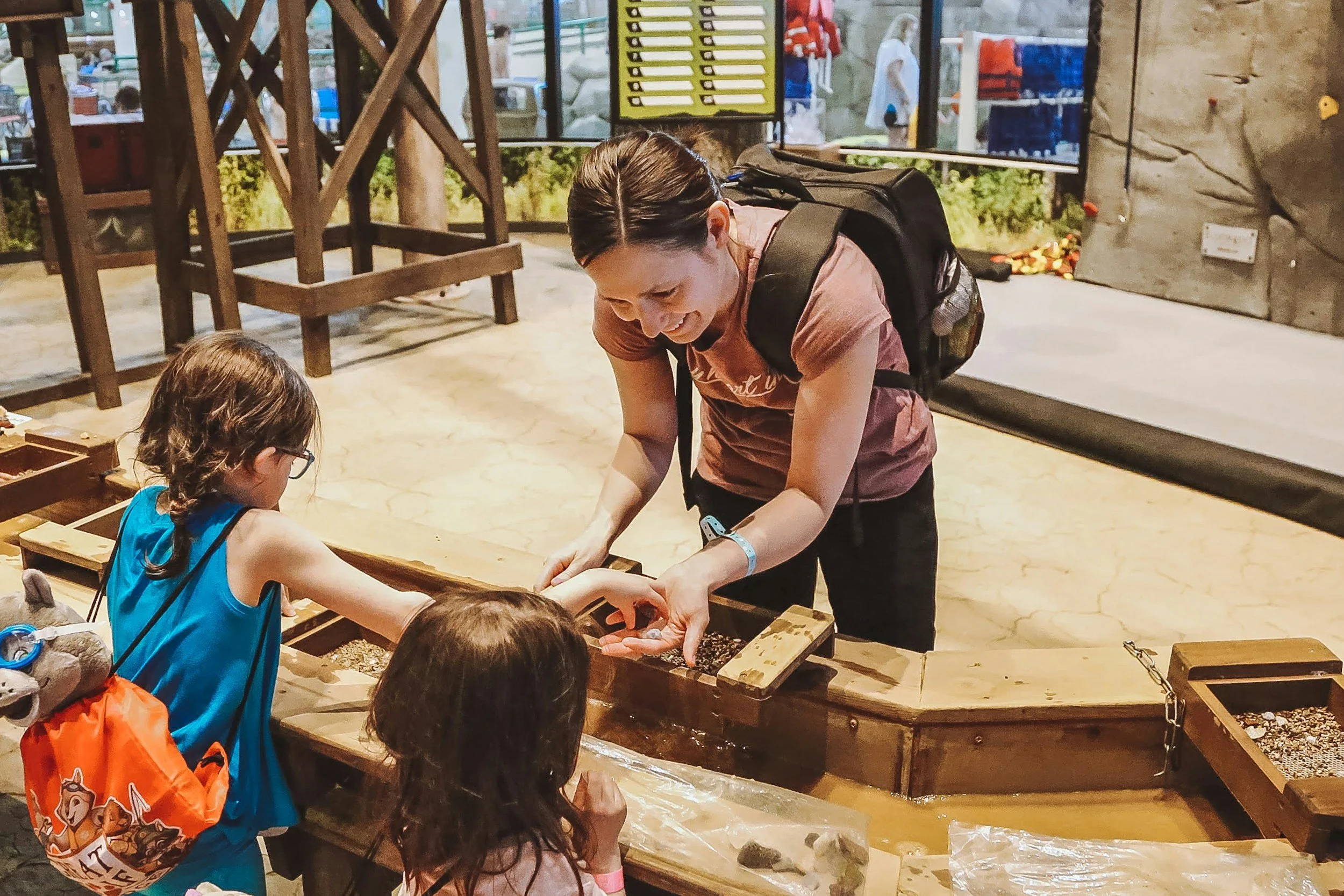 A woman with brown hair in a ponytail and wearing a pink shirt is helping children at a sand and gravel activity station. The children are wearing bright clothing, including a blue top with an orange bag, and are engaged in the activity. The background includes a large window, wooden chairs, and a rock climbing wall.
