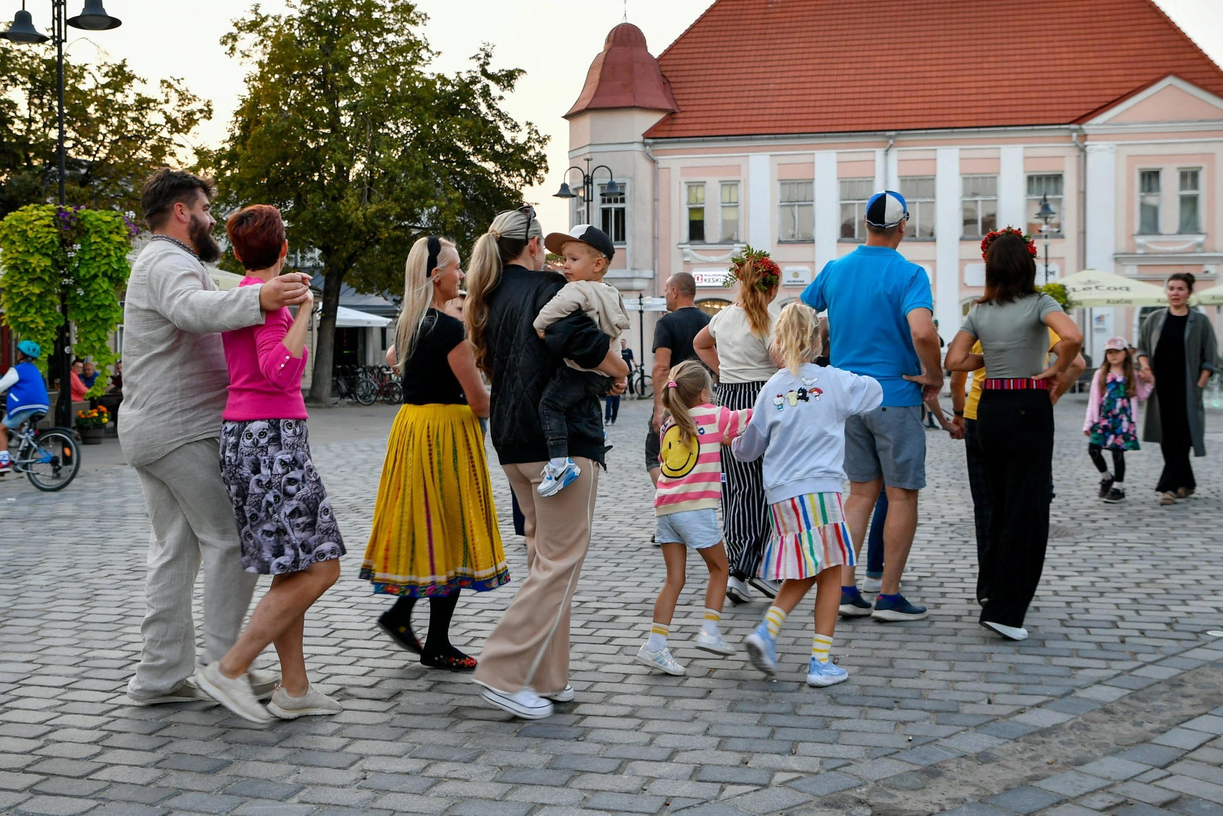 A group of people holding hands and dancing in a circle in a town square with cobblestone pavement, surrounded by trees and historic buildings, during daylight.