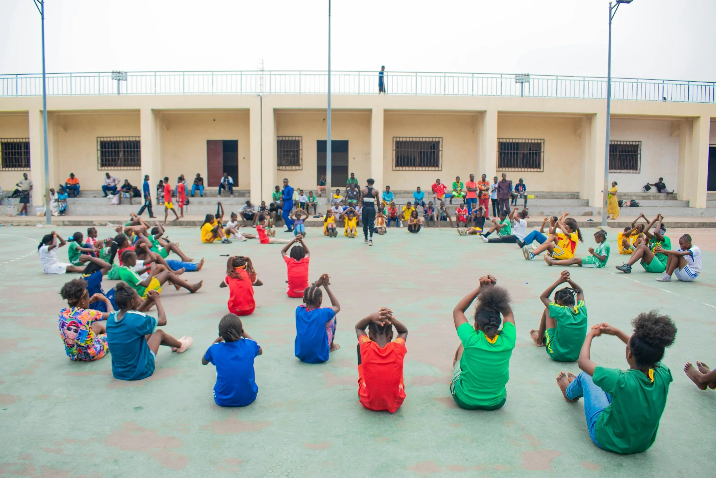 Children sitting on the ground in colorful clothing during outdoor activity, with mostly adults and a few children standing or sitting on benches next to a building, under a partly cloudy sky.