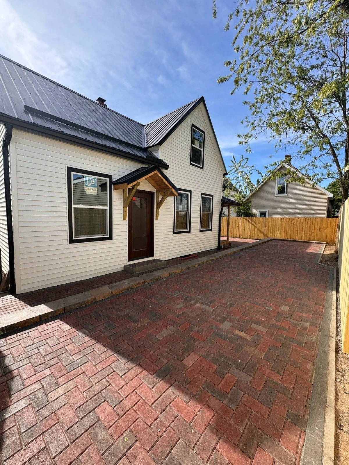 After: side view of the finished home along a brick-paver courtyard, with a timber-framed entry and dark-trimmed windows.