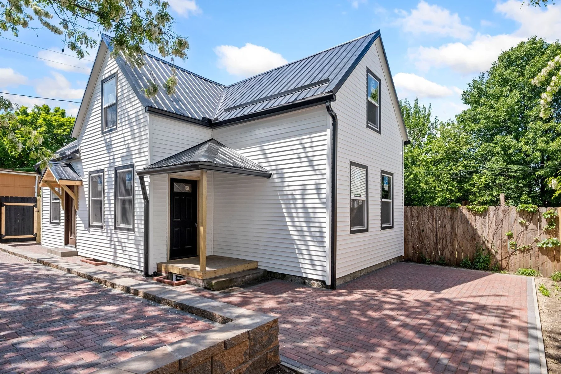 After: the same gabled home finished with light siding, a dark metal roof, black-trimmed windows and a new covered entry with wood posts.